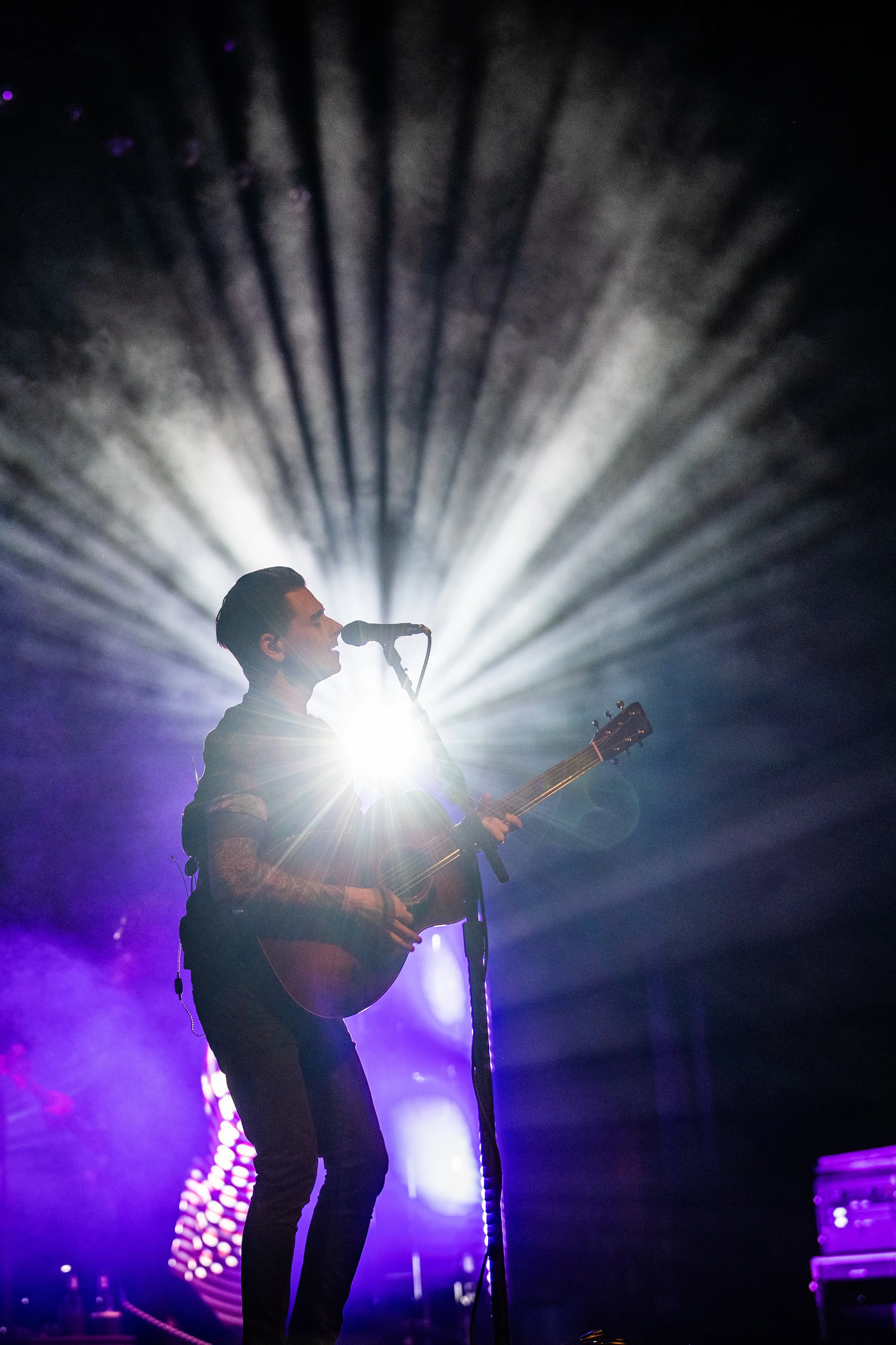 A male musician playing an acoustic guitar and singing into a microphone on stage with bright stage lights shining behind him.