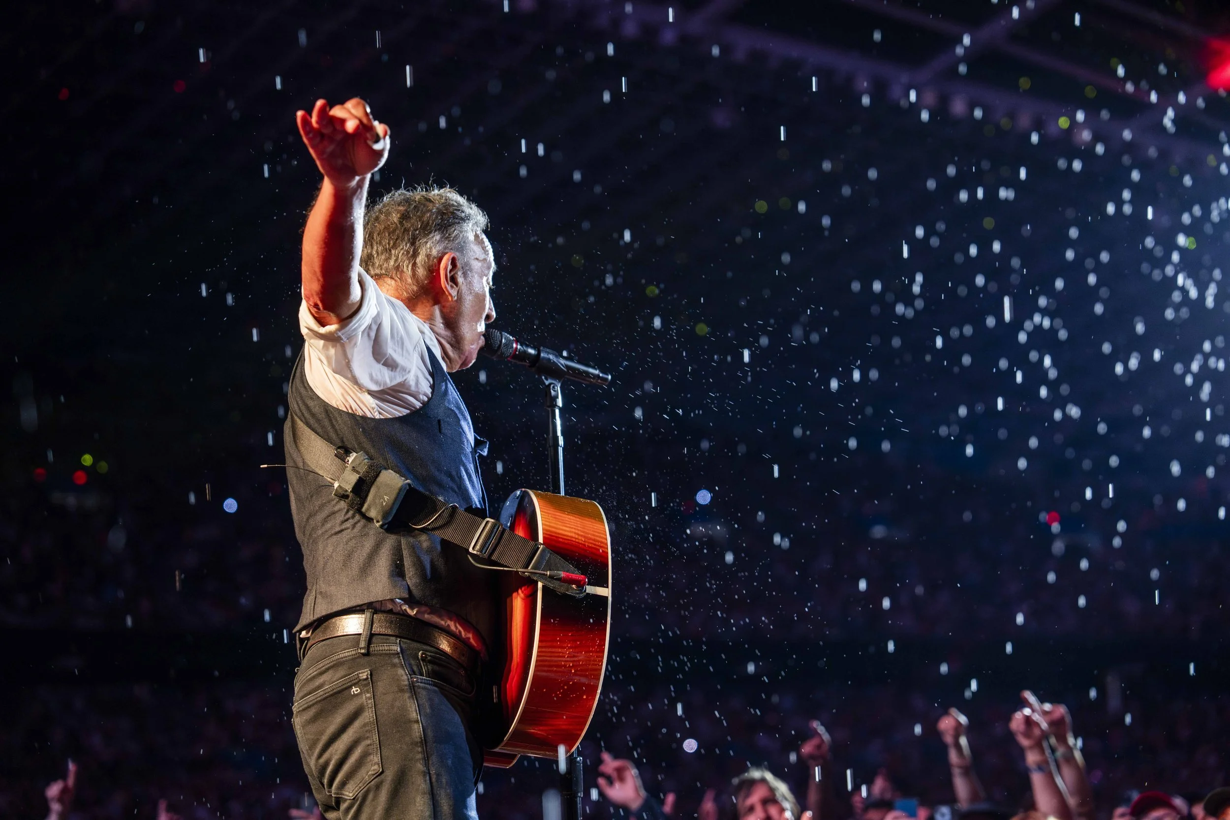 A man playing guitar and singing into a microphone at a concert, with rain or water droplets falling around him and a crowd of fans with raised hands in the background.