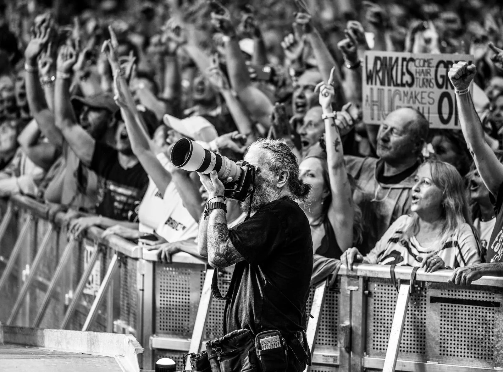 A black and white photo of a crowd at a protest or rally, with many people raising their fists. In the foreground, a photographer with tattoos and a camera with a large lens is taking pictures. Some signs are visible, one mentioning wrinkles and hair turns.