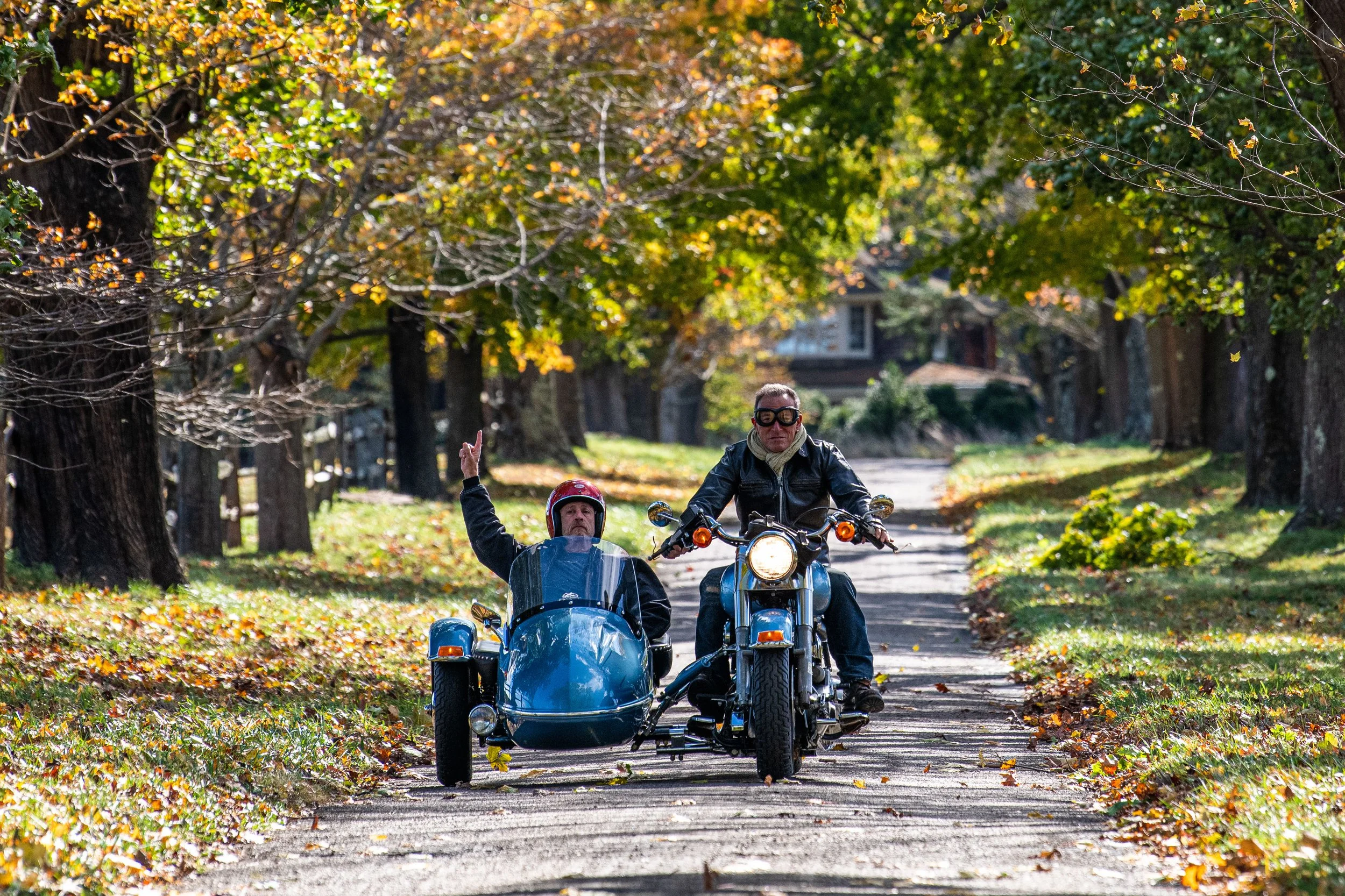 A man riding a motorcycle with a sidecar and a person sitting in the sidecar, waving, on a fall day with colorful trees lining a dirt path.
