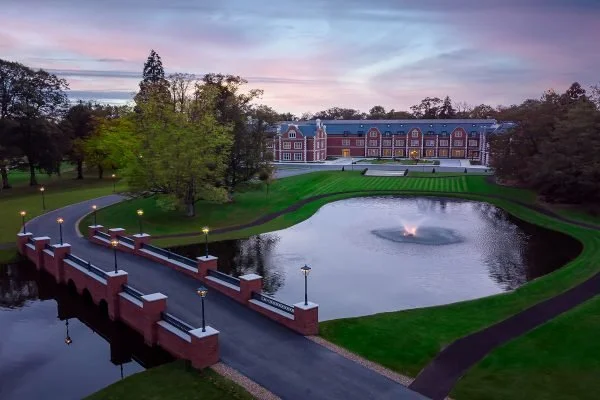 View of a large building in the background with surrounding trees, a pond with a fountain in the foreground, a curved pathway, and a red brick bridge over the pond, all captured during sunset.