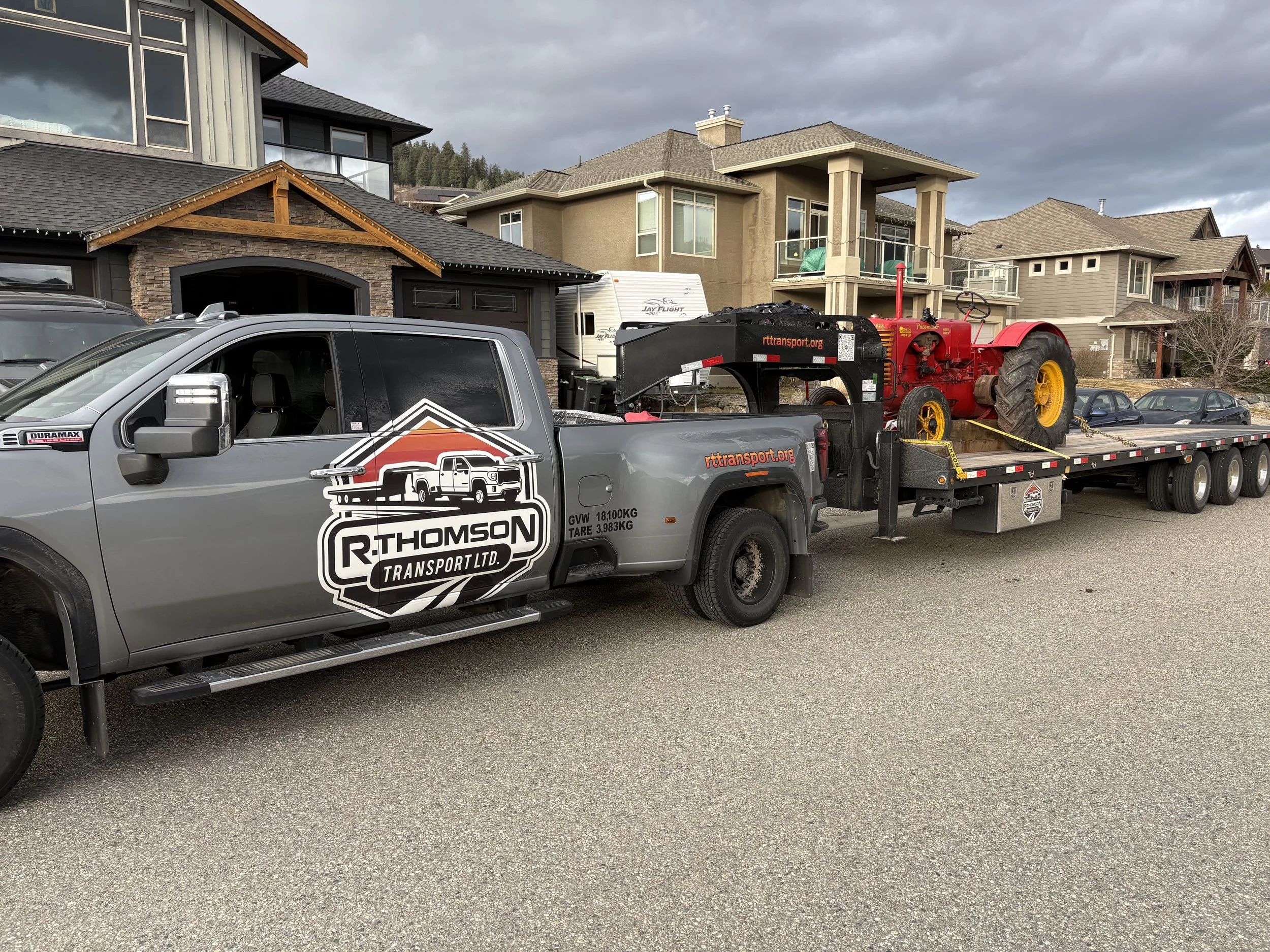 R. Thomson Transport Ltd. GMC heavy-duty dually truck hauling a vintage tractor on a gooseneck flat deck trailer in a residential area in Alberta.
