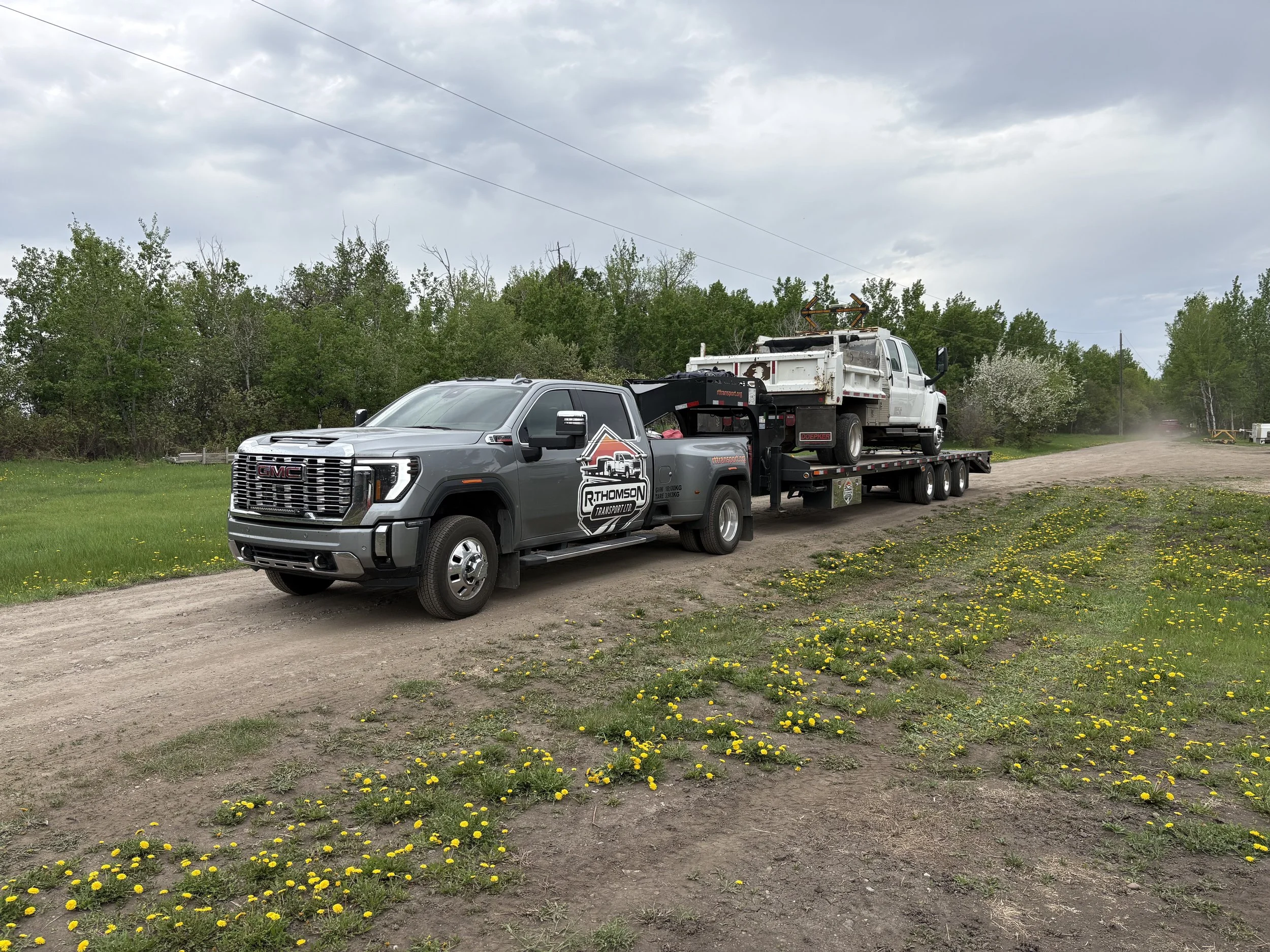 R. Thomson Transport Ltd. GMC heavy-duty dually truck hauling a utility service truck on a gooseneck flat deck trailer along a rural road in Alberta.