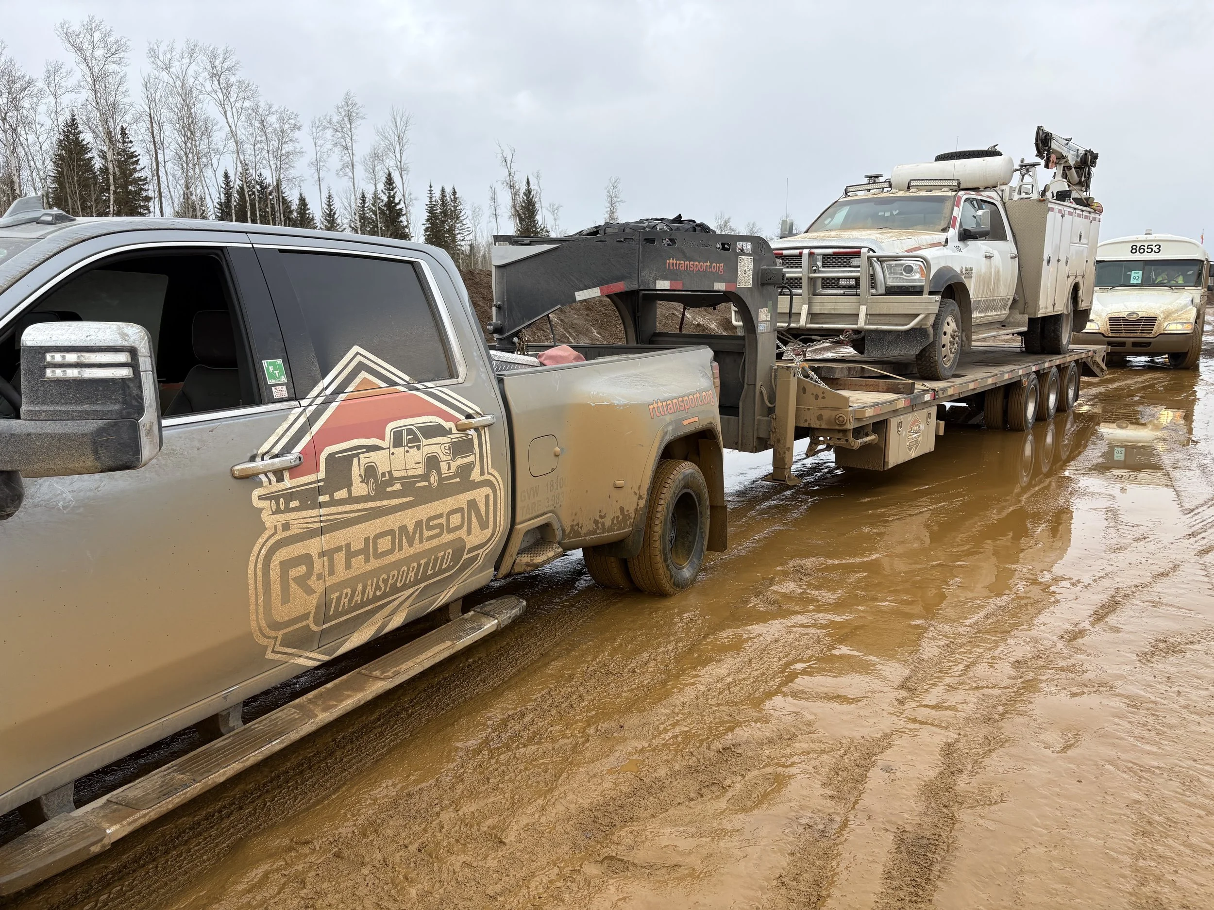 R. Thomson Transport Ltd. GMC heavy-duty dually truck hauling a utility service truck on a gooseneck flat deck trailer through muddy industrial jobsite conditions in Alberta.