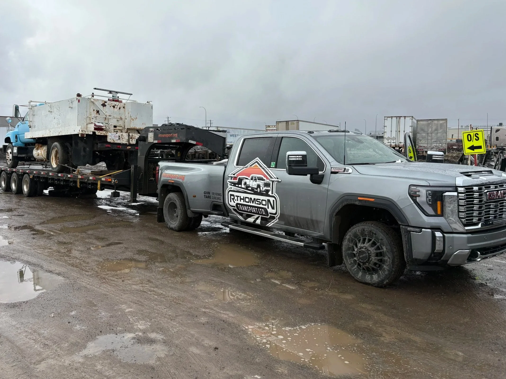 R. Thomson Transport Ltd. GMC heavy-duty dually truck hauling a utility service truck on a gooseneck flat deck trailer at an industrial jobsite in Alberta.