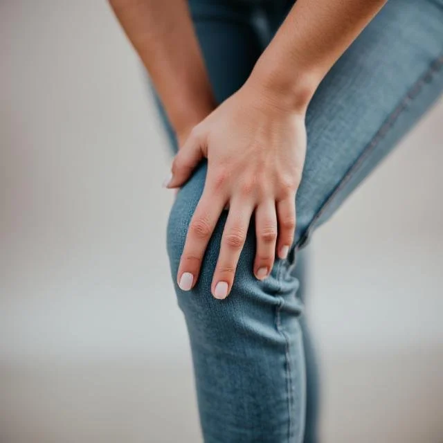 A person with dark skin wearing a black athletic tank top, holding their shoulder with their hand. The background is dark and the lighting highlights the person's shoulder and hand.