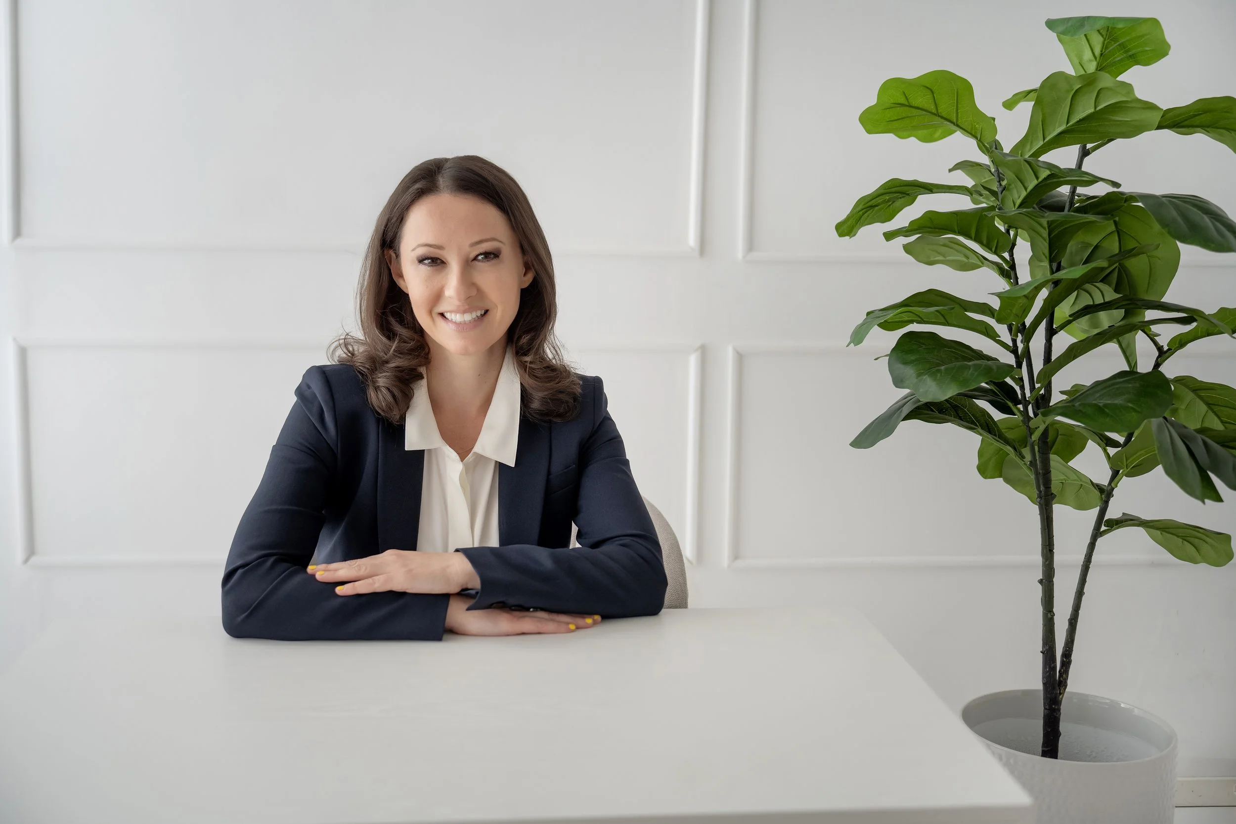 A woman in business attire sitting at a white table, smiling, with a large green plant on the right side of the image in a white pot, and a white wall background.