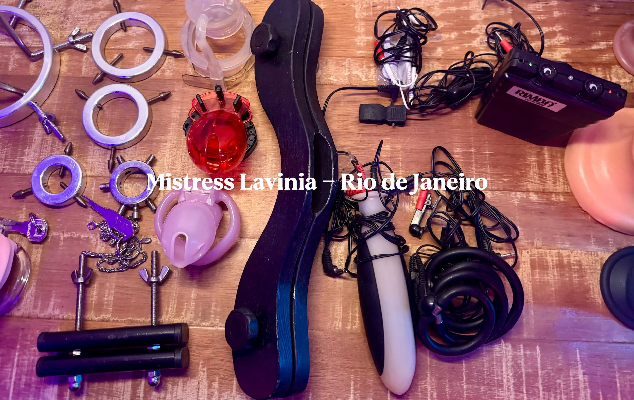 Diversos objetos de BDSM e acessórios de couro e metal, incluindo algemas, chicotes, plugs, cordas, e um astral, dispostos sobre uma mesa de madeira. Texto sobreposto: 'Mistress Lavinia - Rio de Janeiro'.