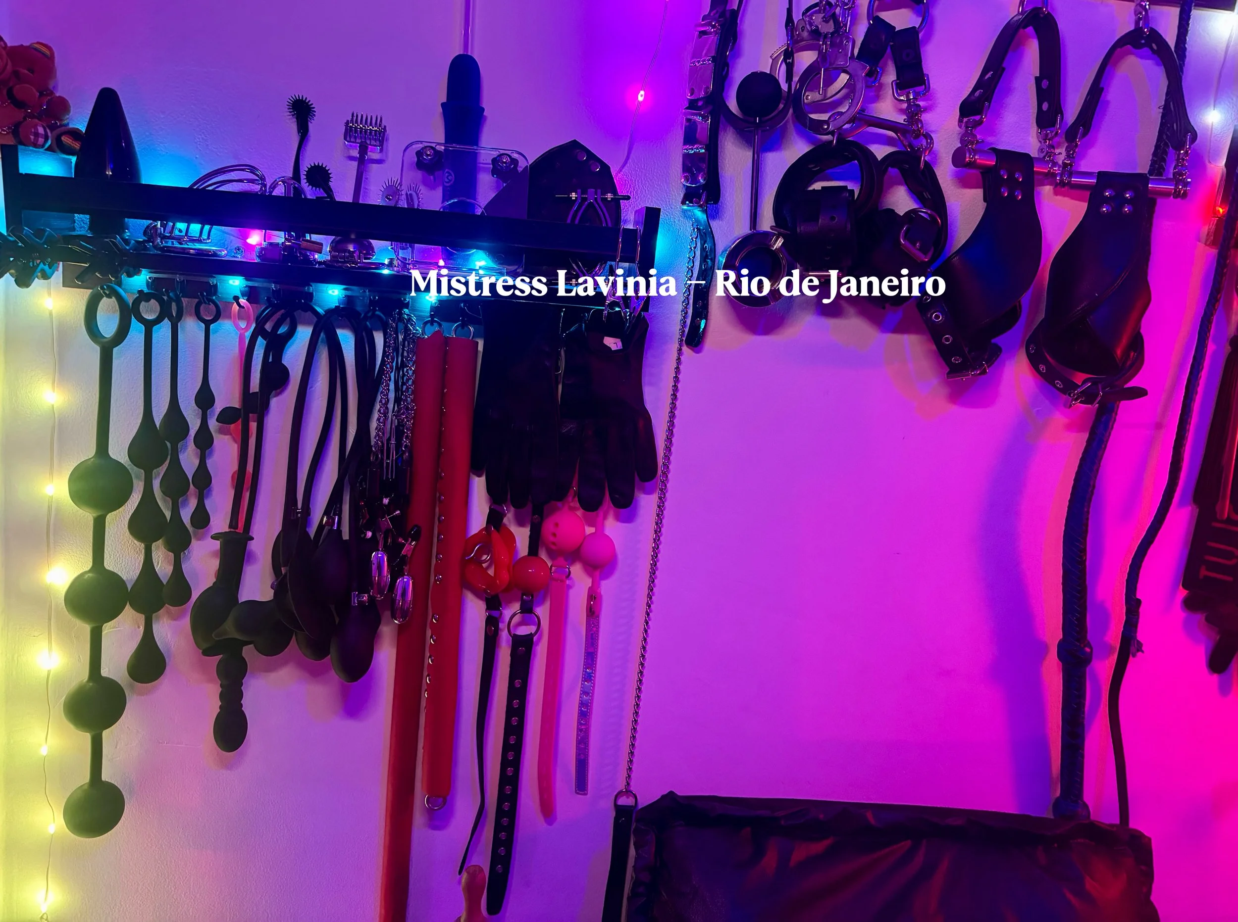 Coleção de acessórios BDSM pendurados em suporte e parede, incluindo chicotes, chaves, algemas, colares, cintas, e outros itens, com iluminação colorida.