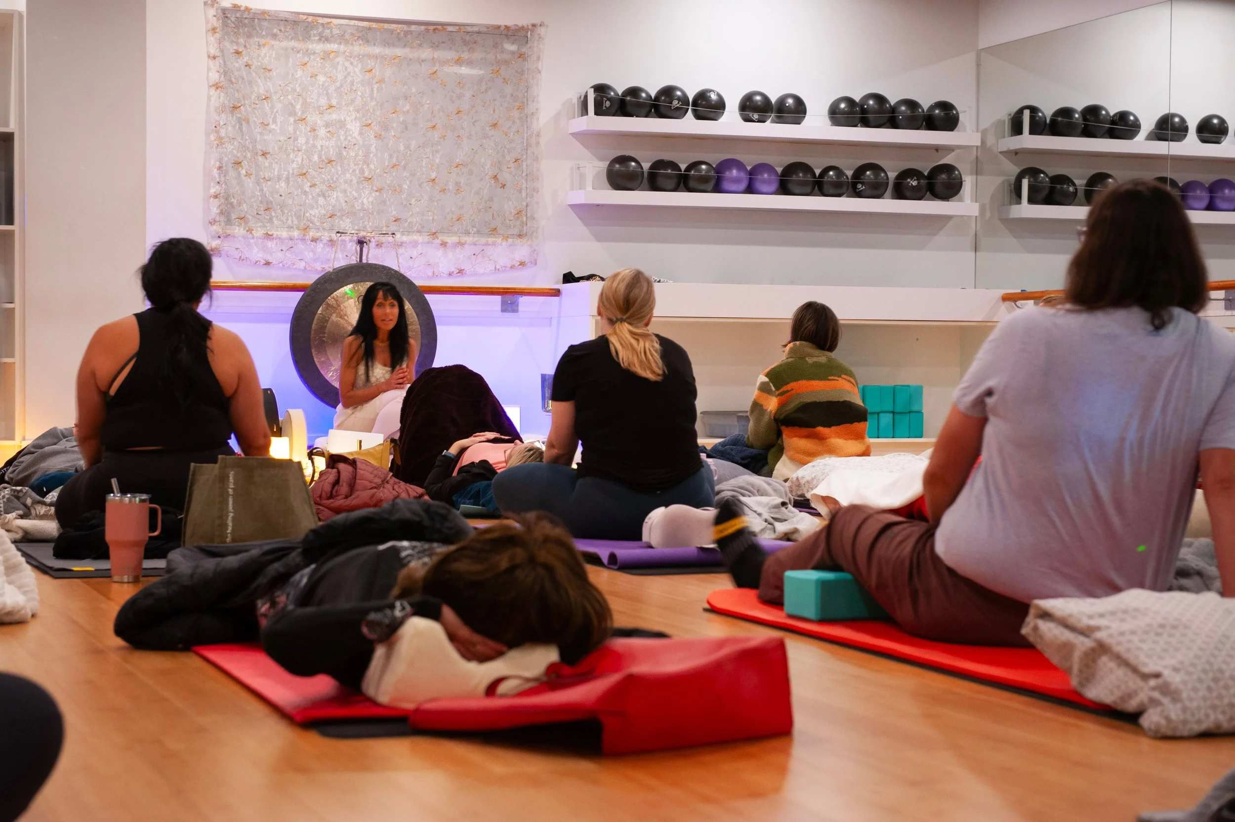 People participating in a yoga class in a studio with mats on the floor, a gong on the wall, and a woman leading the session.