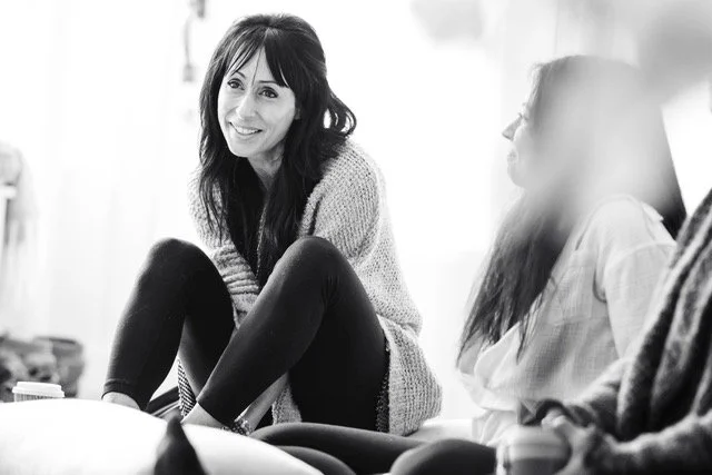 Woman with long dark hair smiling while sitting on a couch with others in a casual setting.