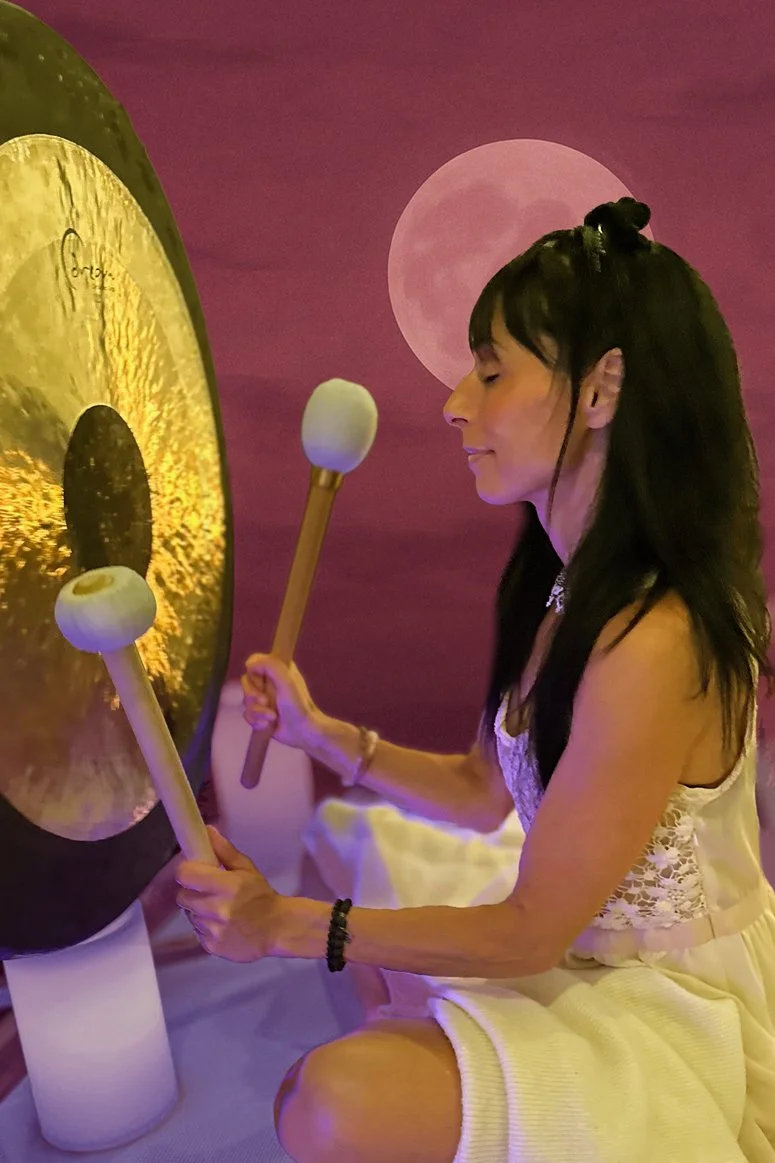 A woman with long dark hair playing a gong with two mallets, a pink moon in the background, and a purple sky.