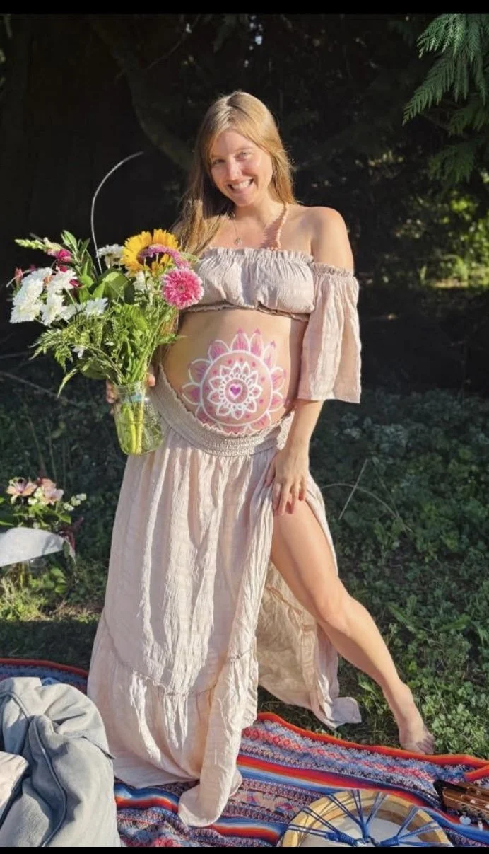 A smiling woman with long blonde hair, holding a bouquet of colorful flowers, standing outdoors in a garden. She has a painted mandala design on her pregnant belly and is wearing a beige crop top and long skirt.