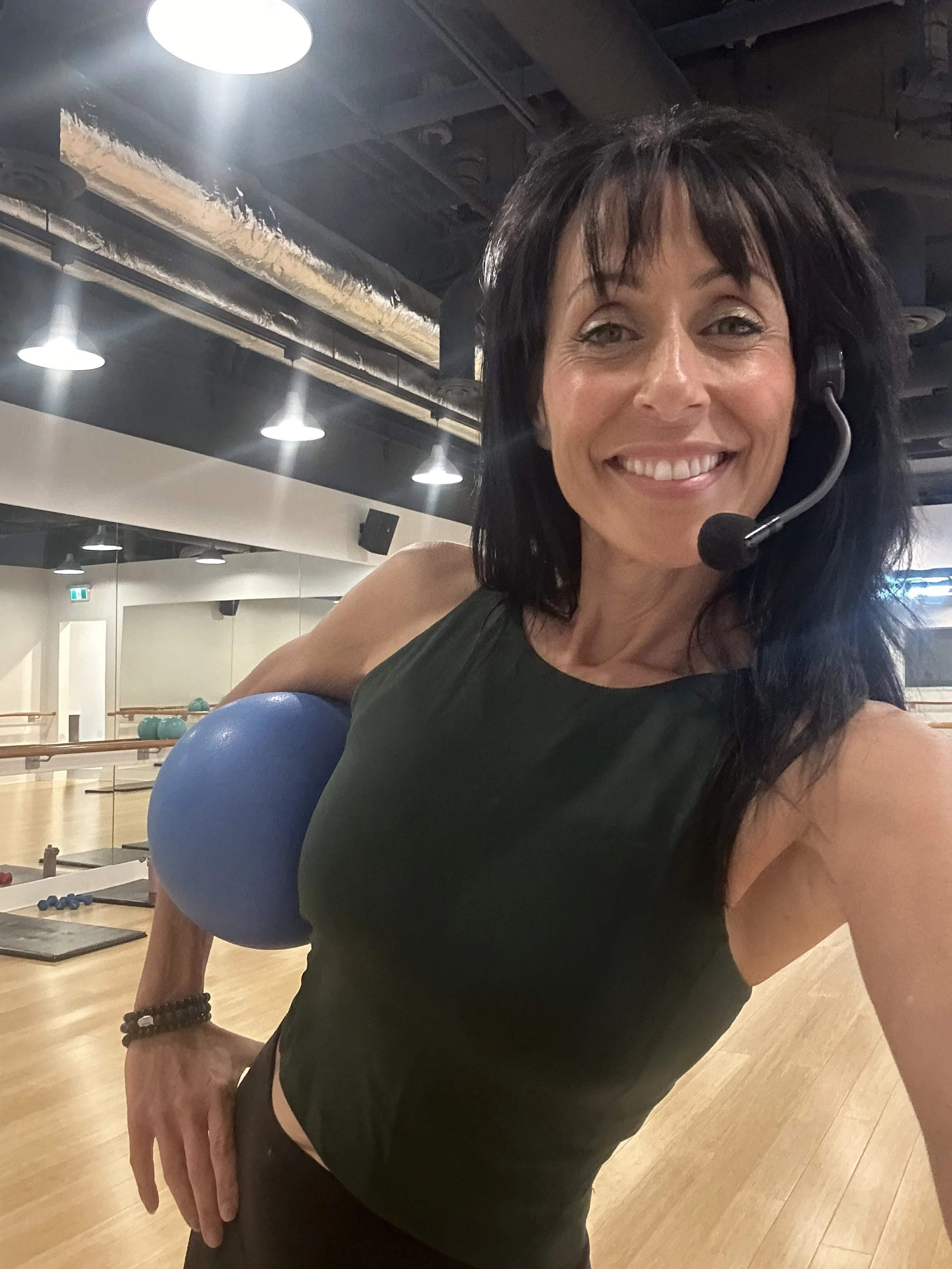A smiling woman with dark hair taking a selfie in a fitness studio. She is holding a blue exercise ball under her left arm and wearing a headset microphone, a dark green sleeveless top, and black workout pants.