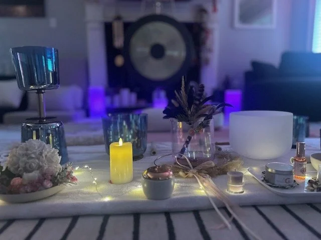 Decorative table setup with candles, flowers, a glass jar with leaves, a white bowl, and various small bottles, with a gong and electronic equipment in the background, illuminated by soft purple lighting.