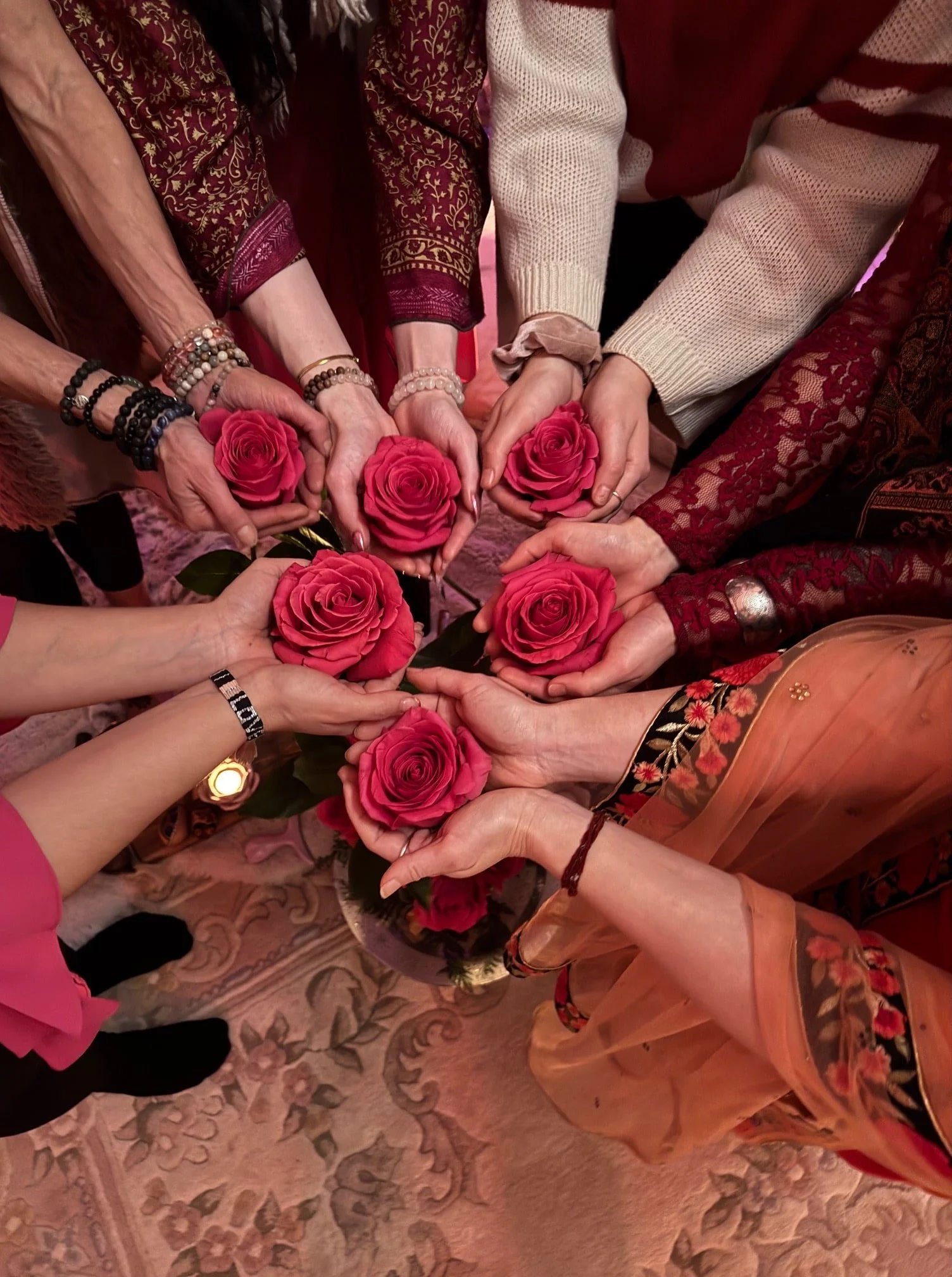 Multiple hands holding pink roses in a circle, with people dressed in colorful, patterned clothing.
