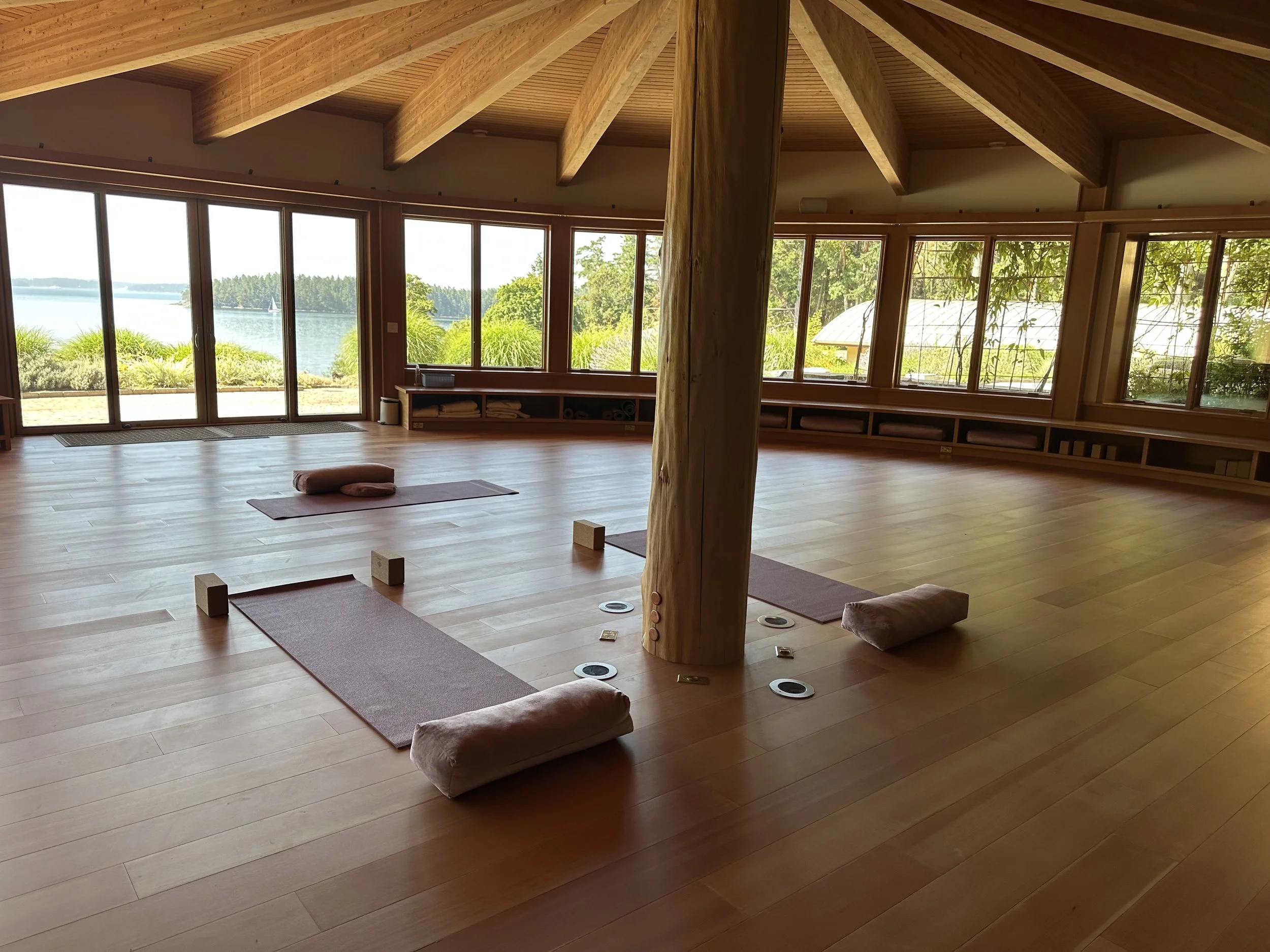 A spacious yoga studio with wooden floors, large windows overlooking a lake and greenery, and yoga mats with bolsters and blocks arranged for a class.