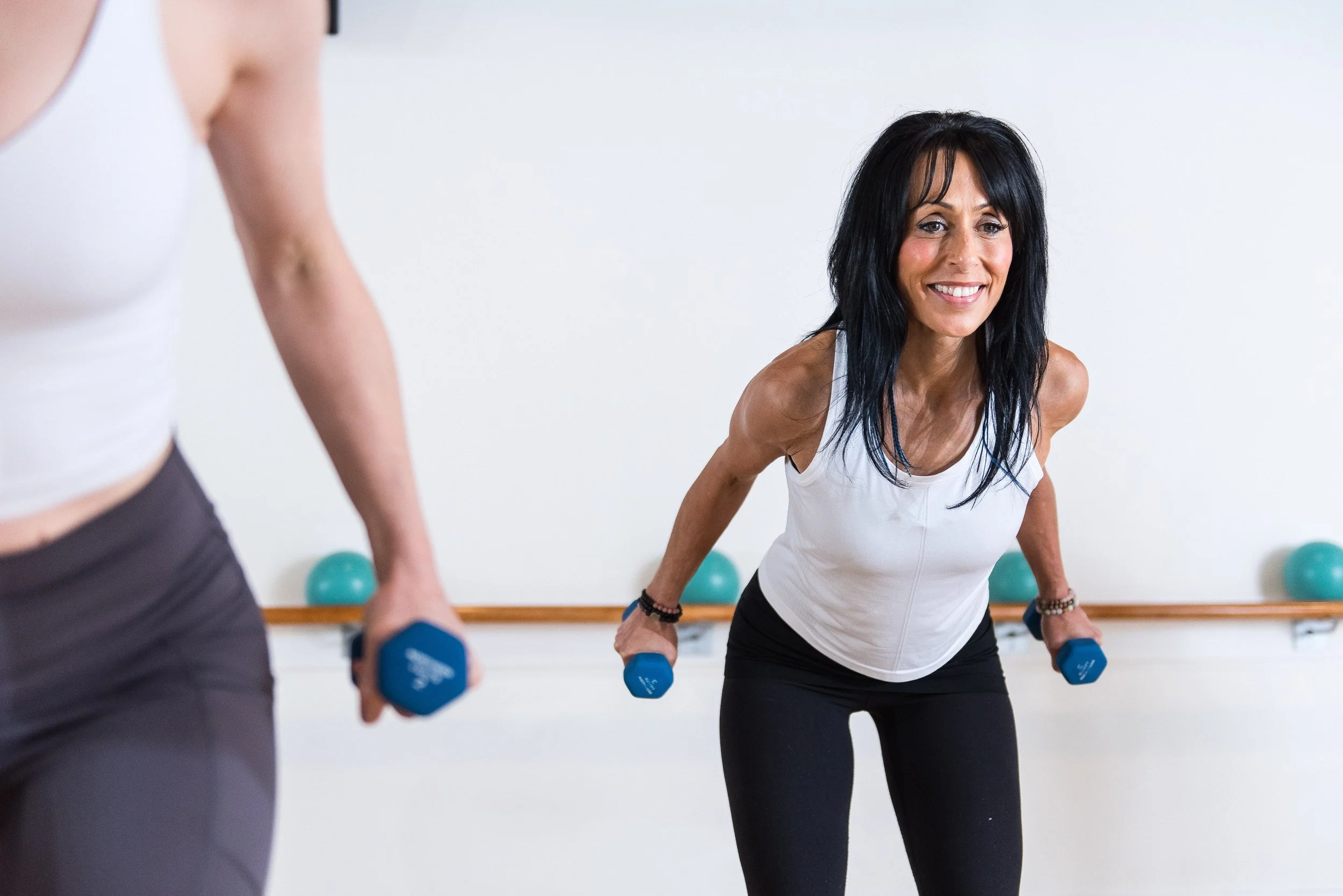 A woman with black hair wearing a white tank top and black pants exercises with dumbbells in a gym.