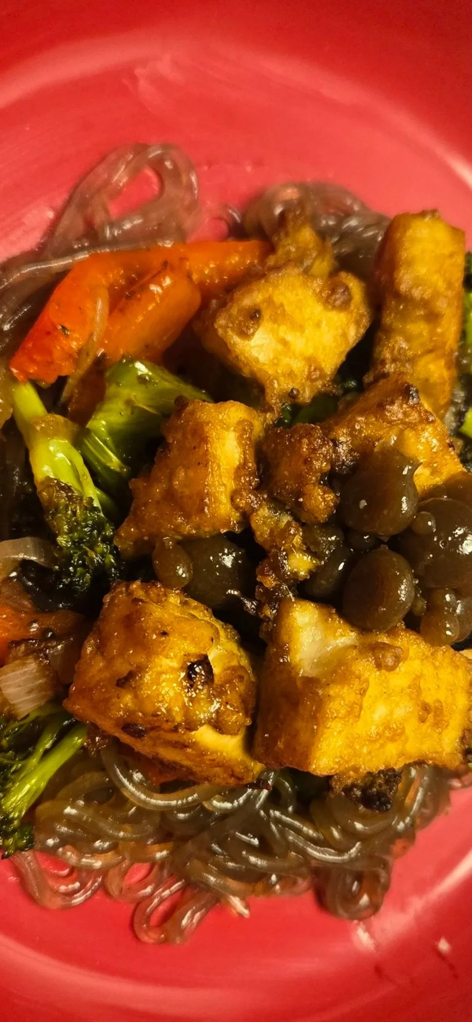 Close-up of a dish with fried tofu, sautéed broccoli and carrots over sweet potato noodles in a pink bowl.