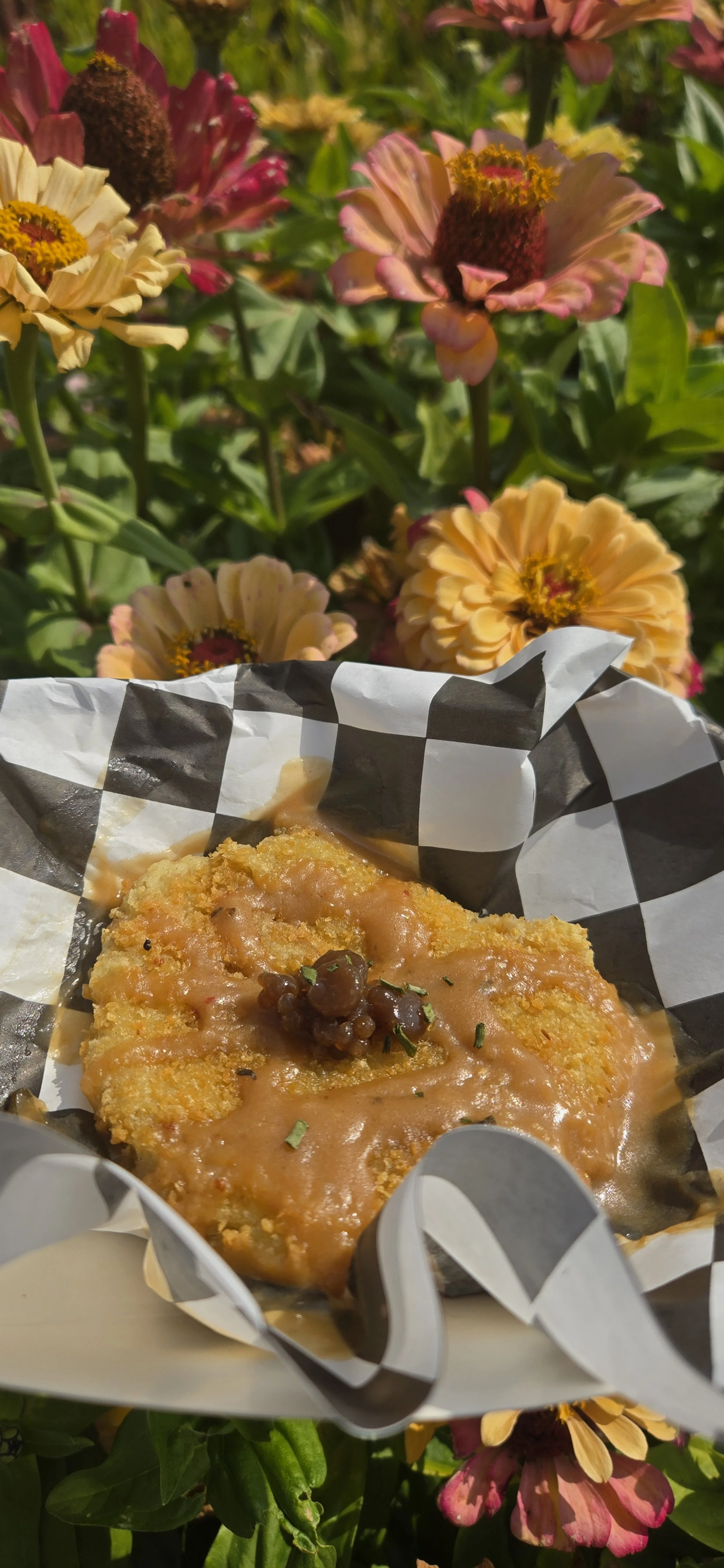 Fried food with gravy topped with a small cluster of black garlic caviar, set against a colorful garden of flowers.