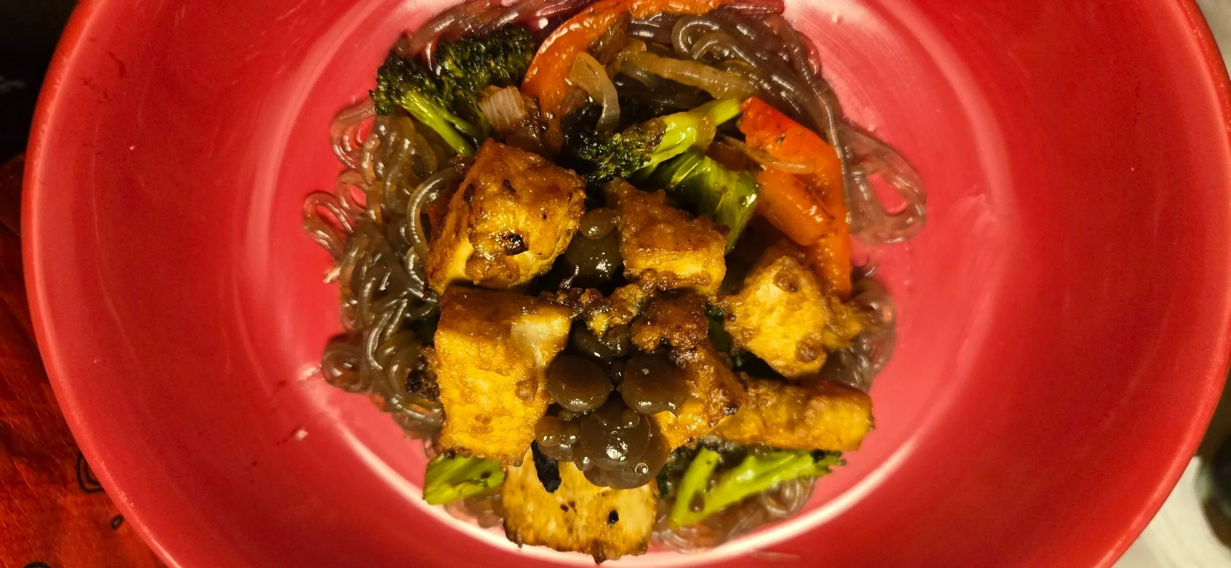 A bowl of cooked black bean noodles topped with stir-fried broccoli, carrots, and tofu cubes.