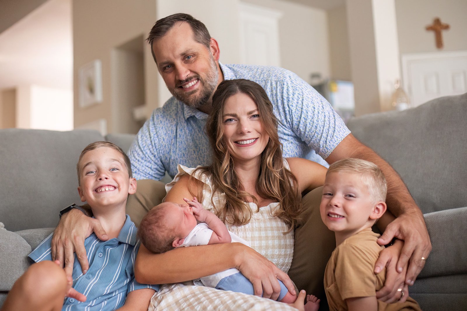 A happy family of five sitting on a couch in a living room, including a man, a woman, two young boys, and a newborn baby.