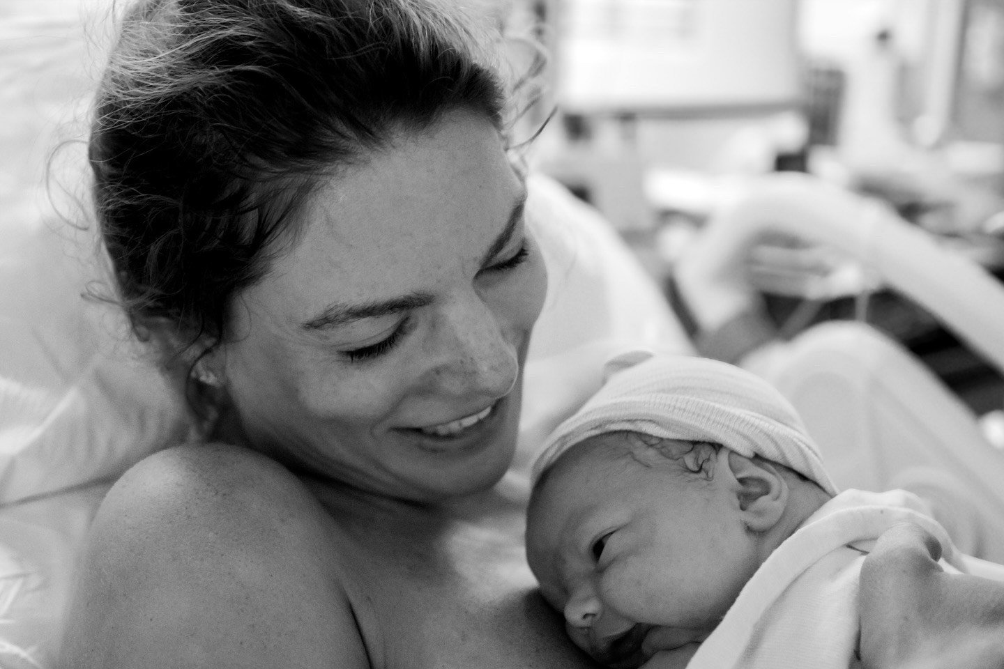 A woman lying in a hospital bed holding a newborn baby, both smiling and looking at each other.