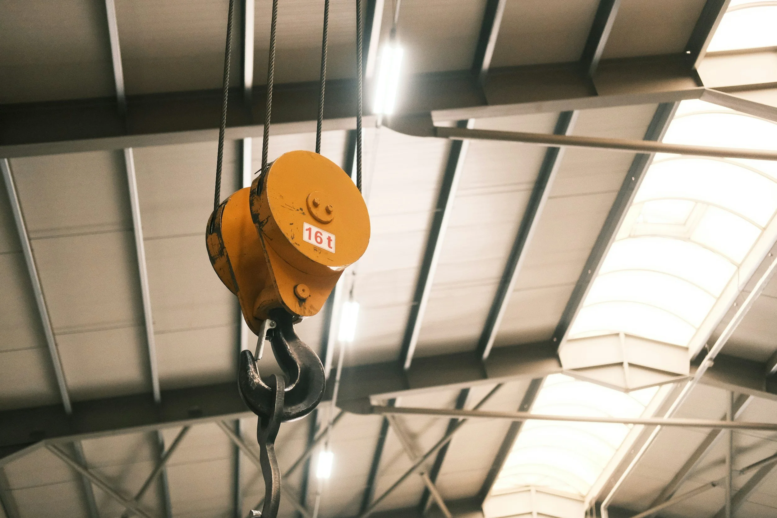 En gul kran med lastkapacitet på 16 ton hänger i taket av ett industriutrymme med takbalkar och en veckad plasttak.