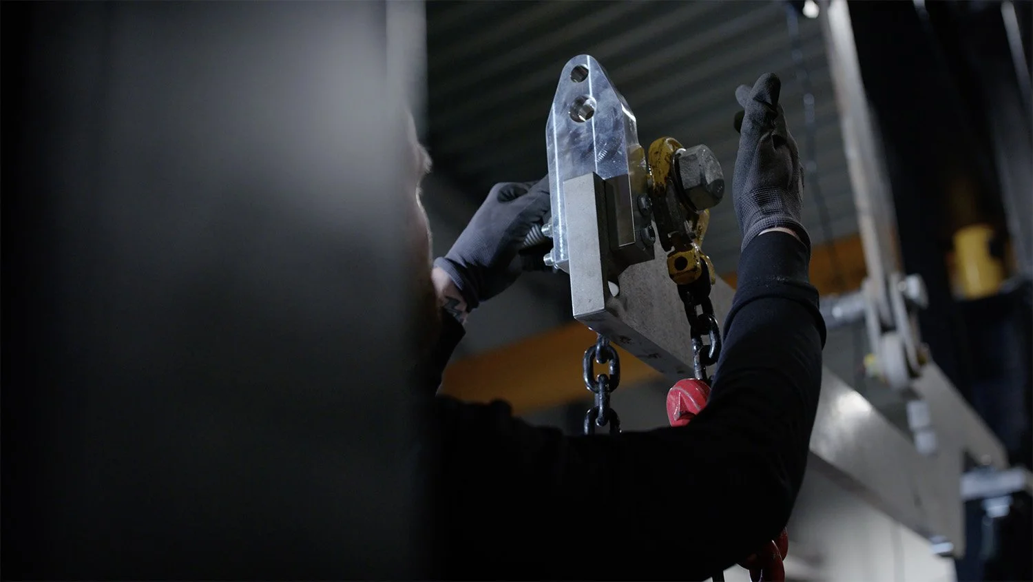 En person i svart overall och handskar arbetar med en metallkomponent i en industrilokal.