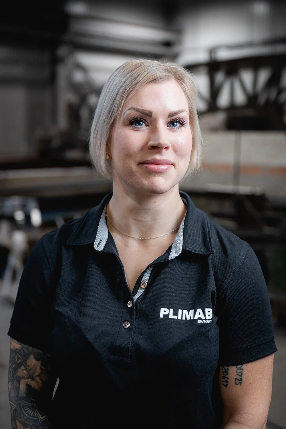 Kvinna med blond hår, klädd i en svart t-shirt med texten 'PLIMAB sweden', står i en industriell miljö.