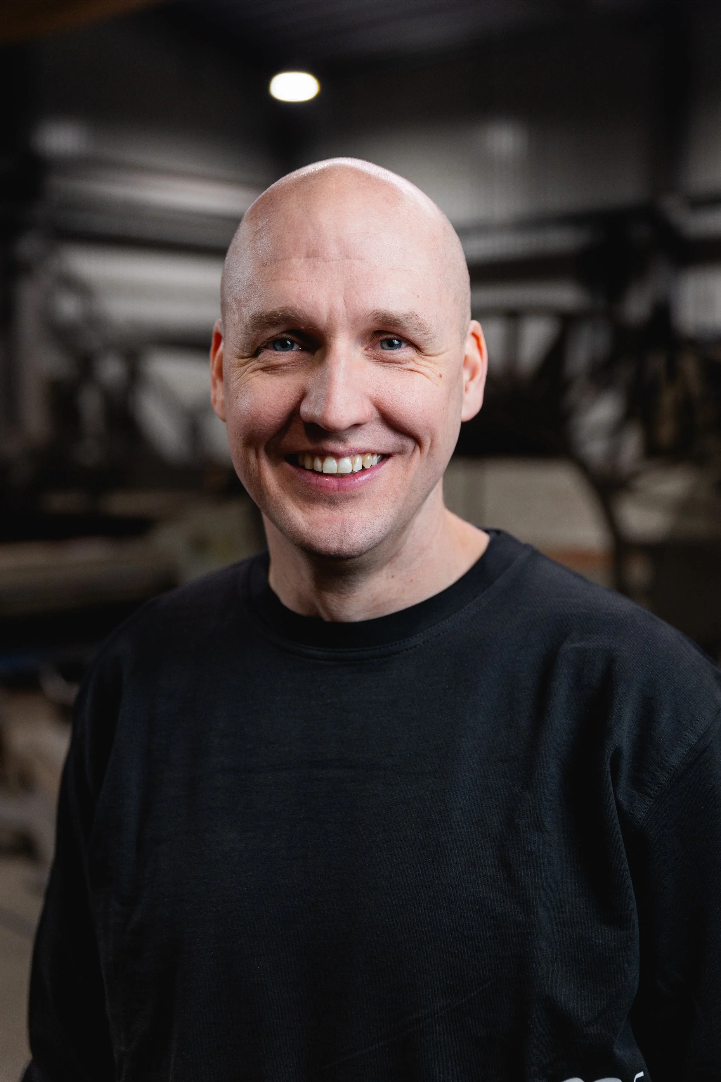 Ung man med rakat huvud, ler och bär svart tröja, står i en industriell miljö.