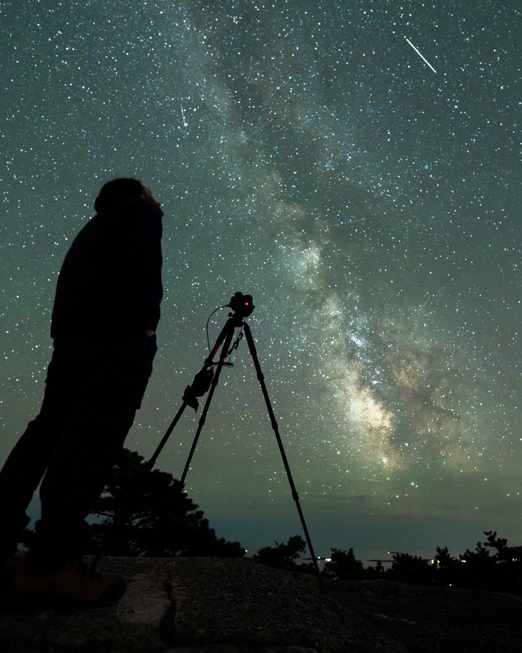 Silhouette of a person standing next to a camera on a tripod, looking up at a starry night sky and the Milky Way galaxy.