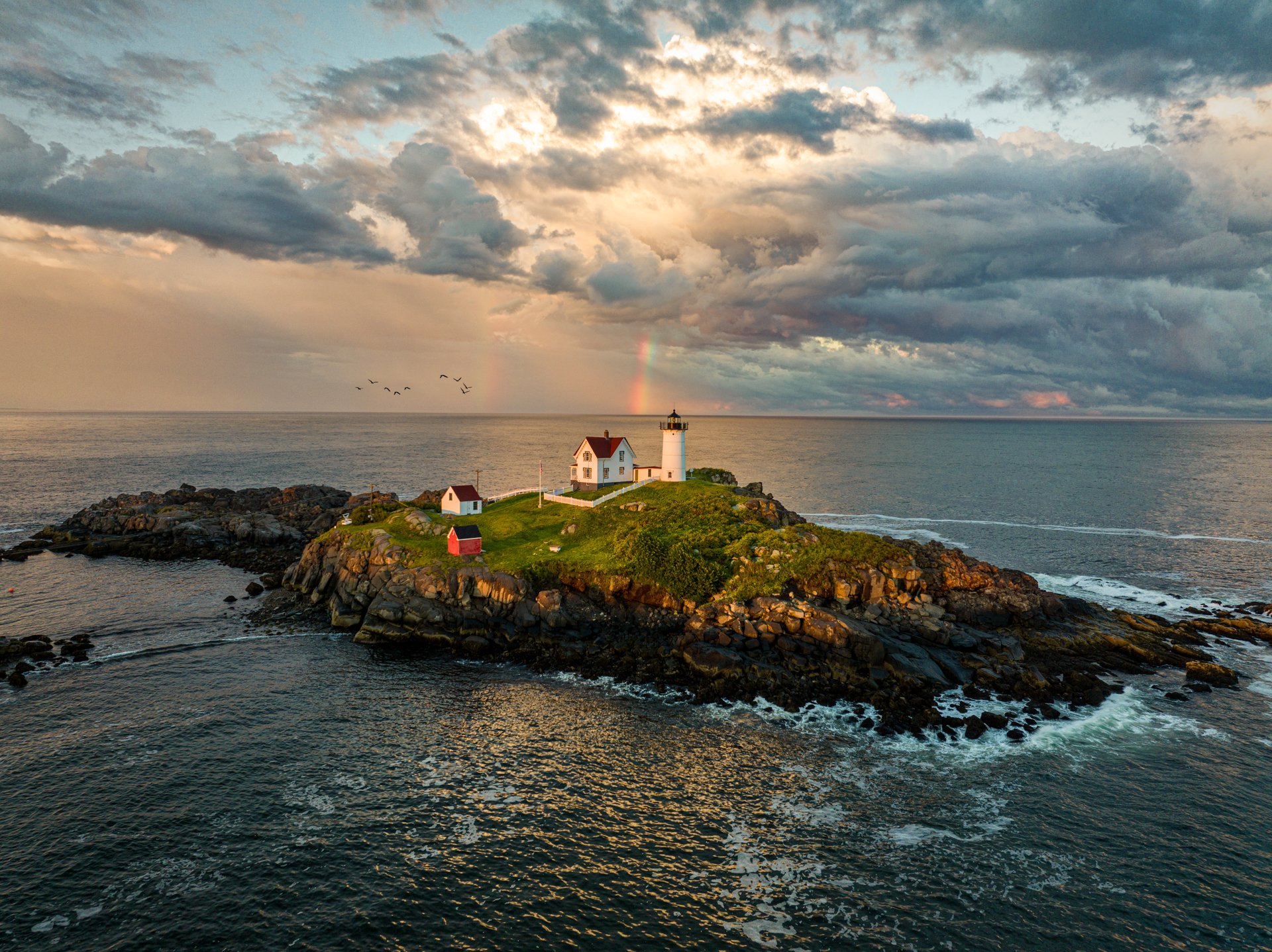 A lighthouse and small houses on a rocky island at sunset with a rainbow in the background over the ocean.