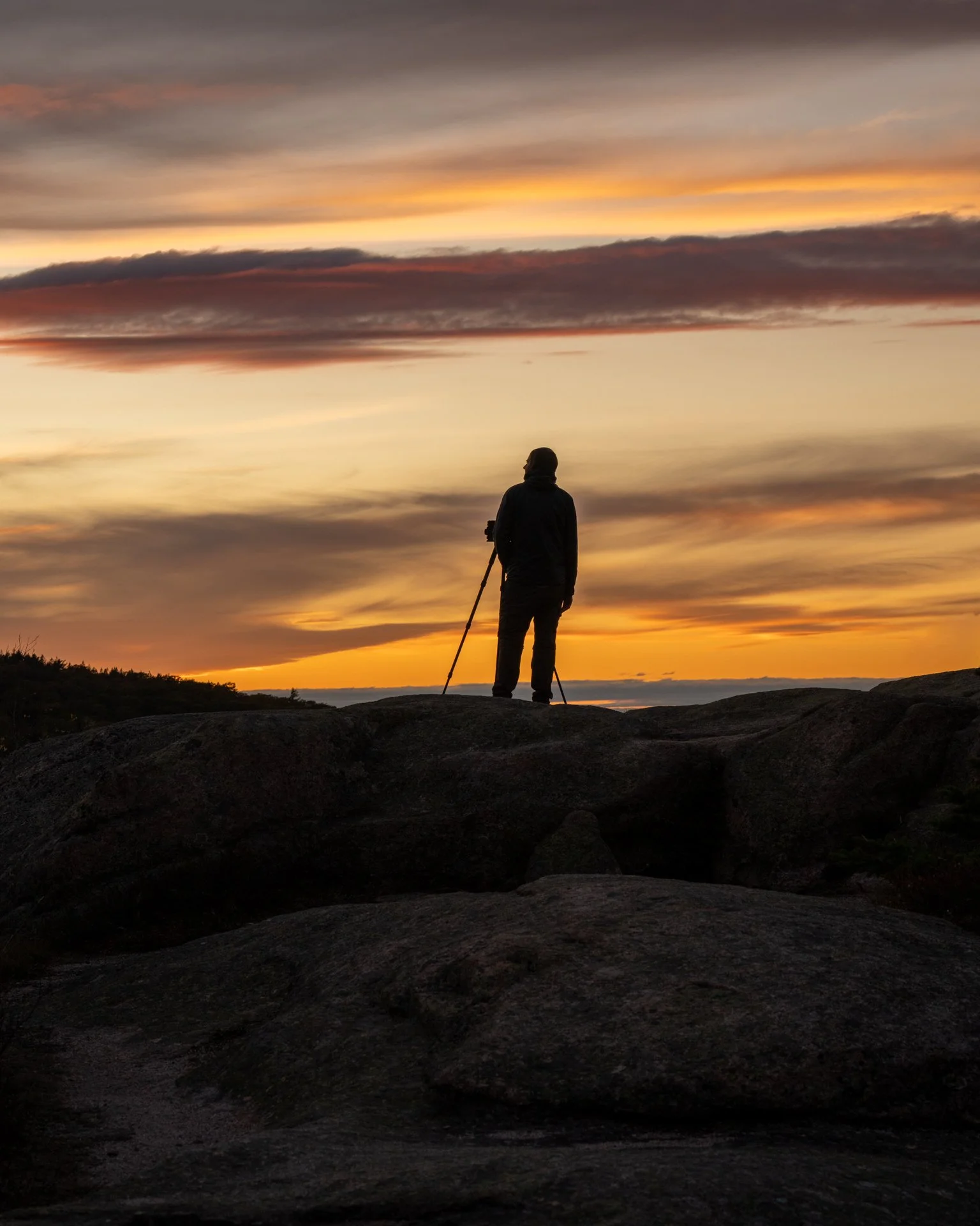 Silhouette of a person standing on rocks and holding a walking stick, watching a colorful sunset sky.