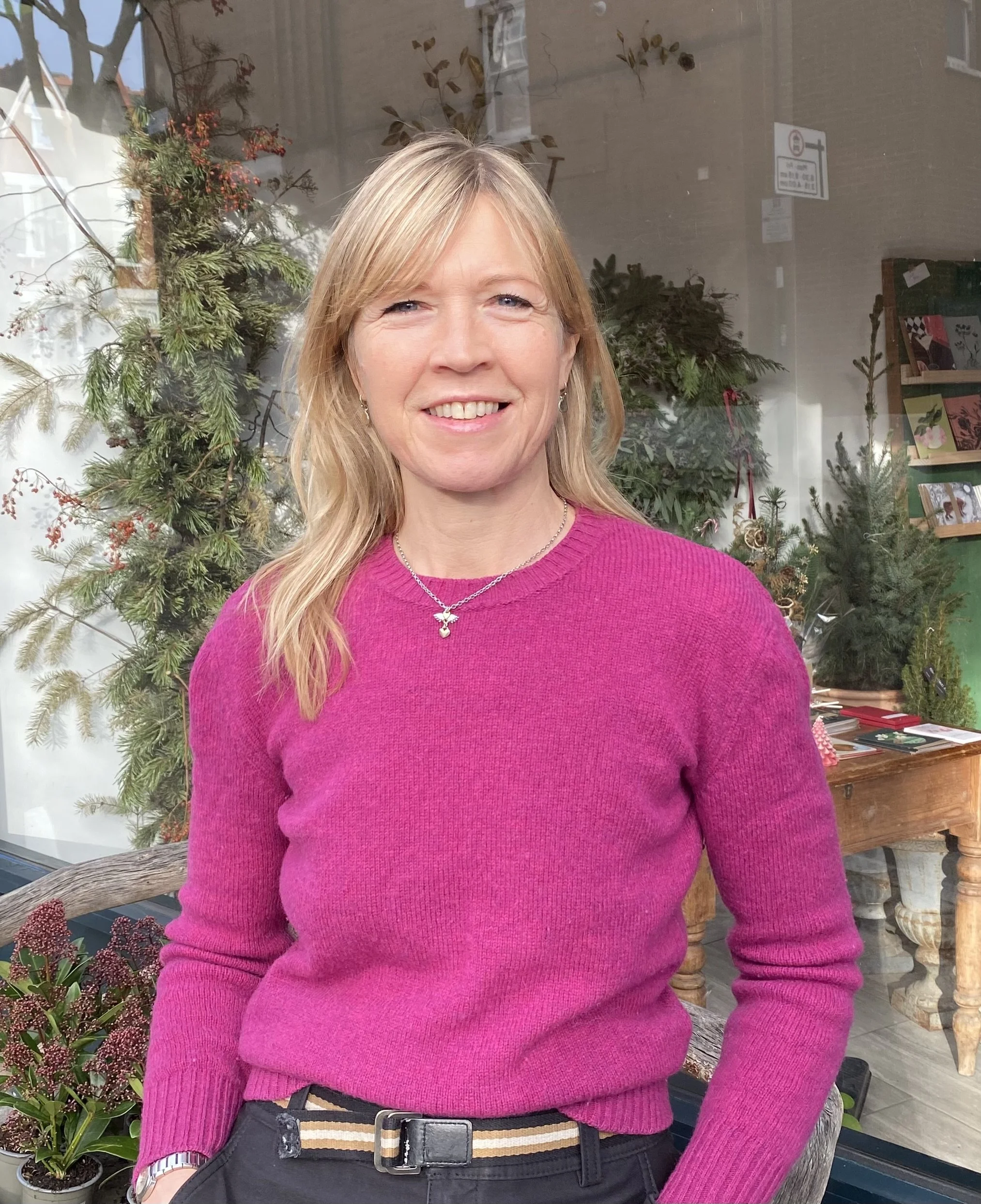 A woman with blonde hair and fair skin wearing a pink sweater, standing outdoors in front of a shop window decorated with plants and decorations.