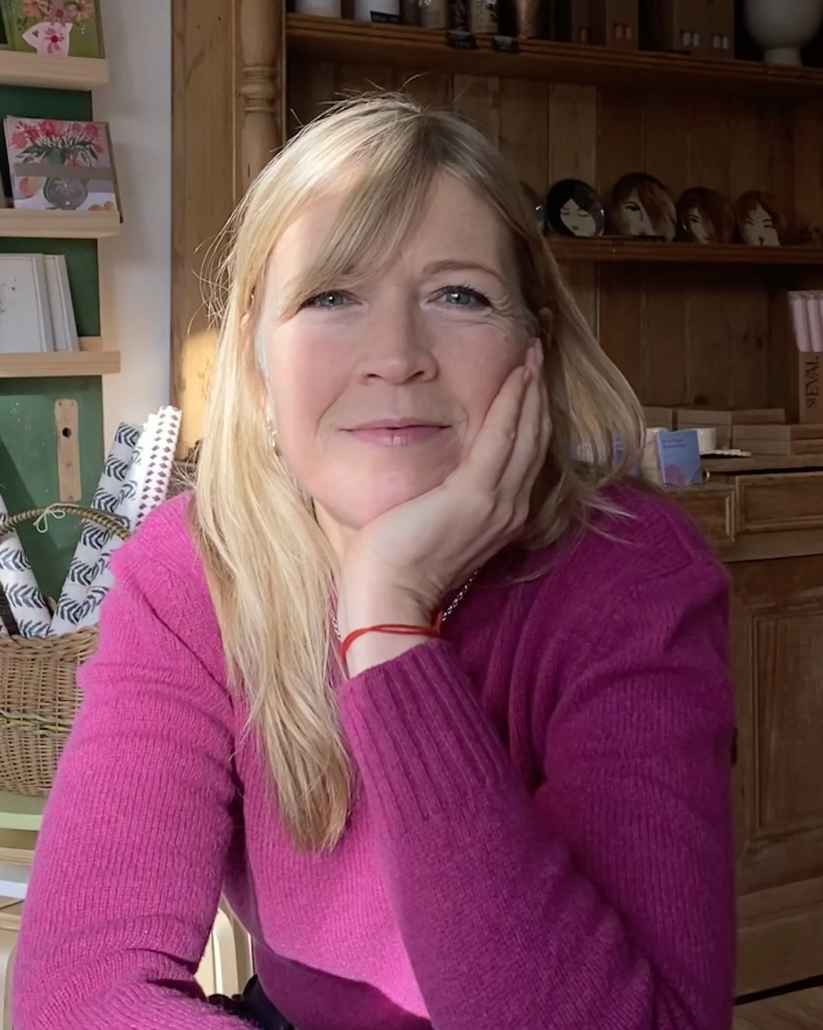 A woman with blonde hair wearing a pink sweater, resting her face on her hand, in a cozy wooden room with shelves and decorative items in the background.