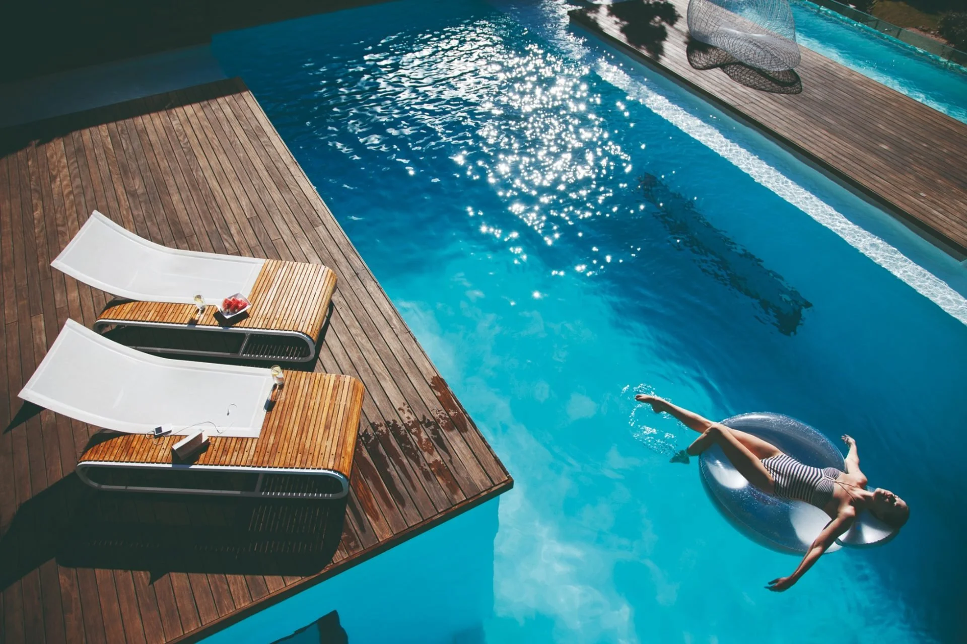 A swimming pool on a wooden deck with three lounge chairs and a small tray with snacks and drinks. A person is floating in the pool on a silver inflatable ring, wearing a striped swimsuit and a gray hat, arm extended.