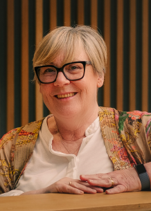 Smiling woman with short blonde hair, glasses, and colorful clothing sitting at a table