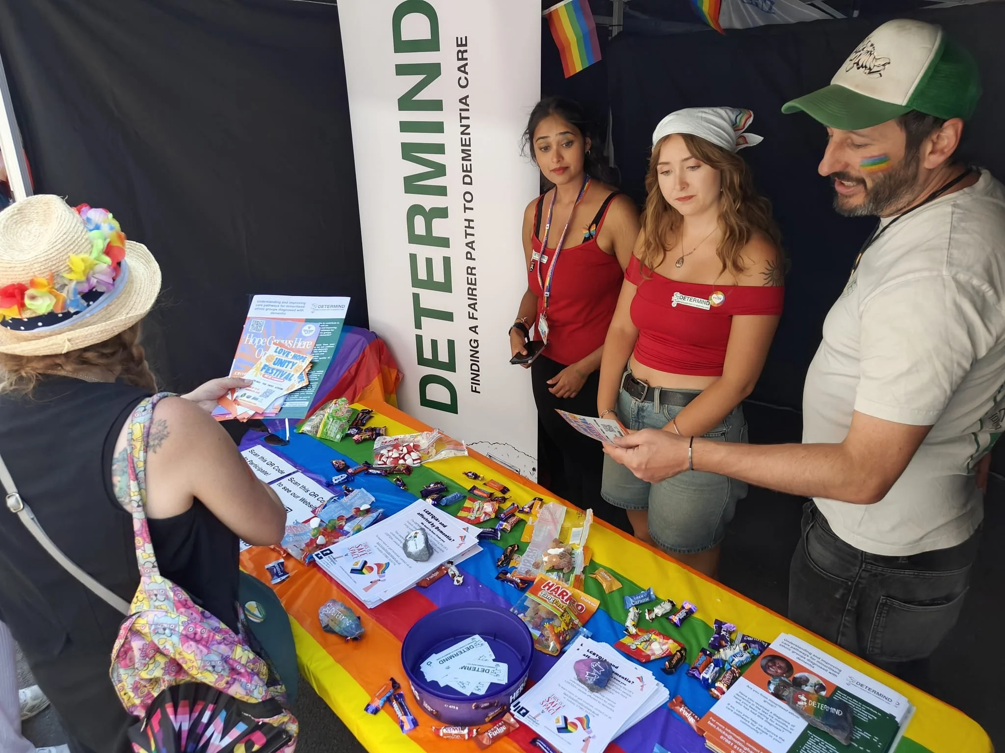 Someone picking up a flyer from the DETERMIND pride stall. The stall has a brightly coloured rainbow tablecloth on it, with lots of sweets for passers by to pick up. There are three of the DETERMIND team standing behind the table smiling.
