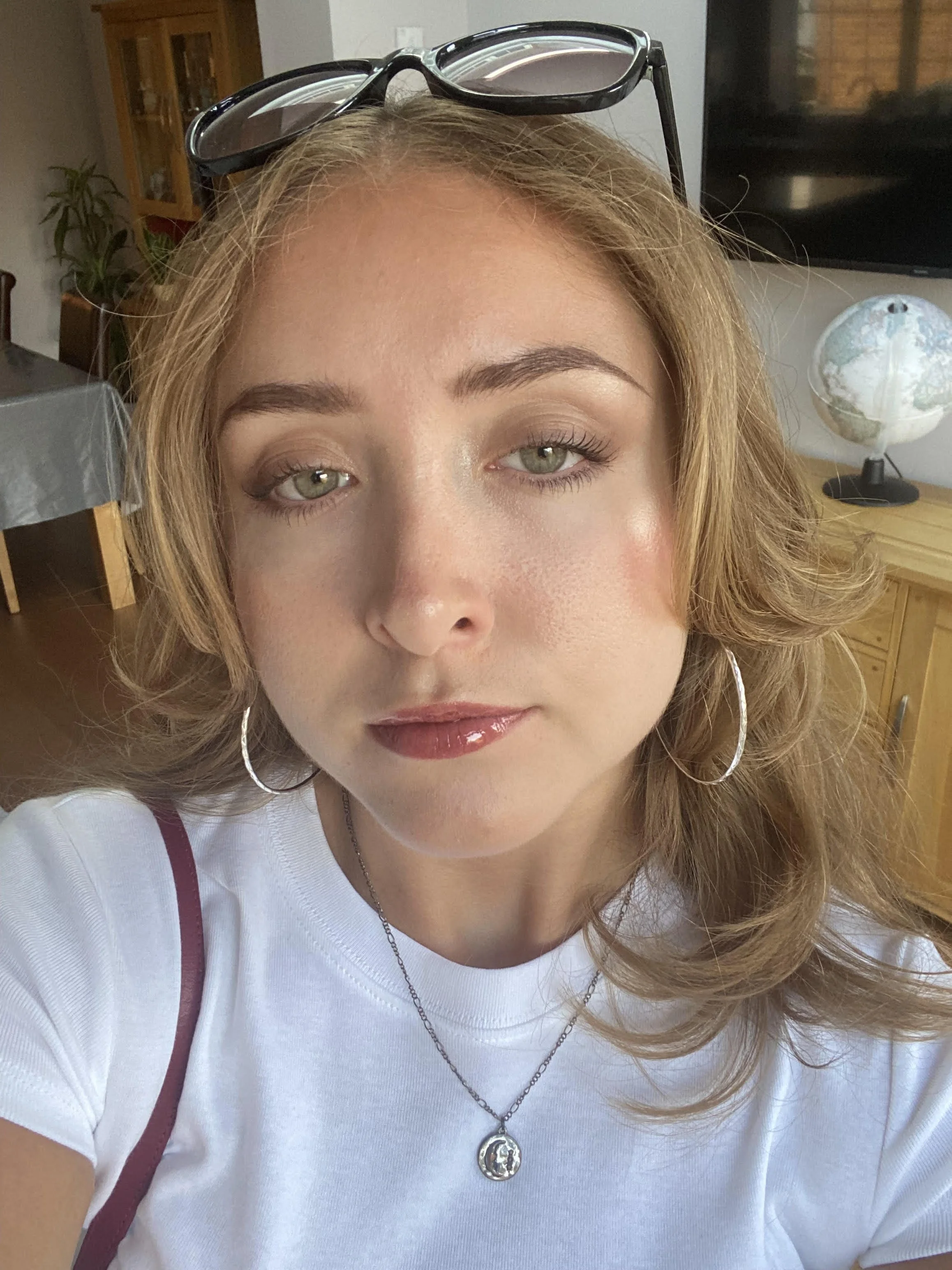 A person with long red hair. She is taking the photo of herself and is wearing a white t-shirt, hoop earrings and has sunglasses balanced on her head.