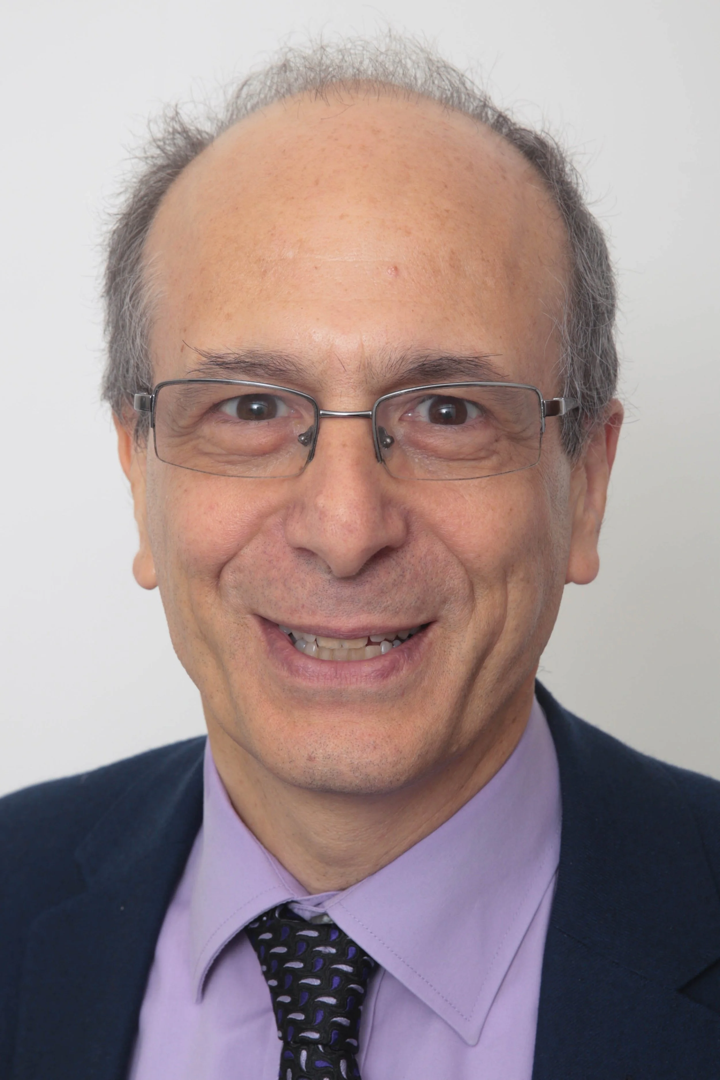 A man with glasses smiling, wearing a lavender shirt, a dark blazer, and a patterned tie, against a plain light background.