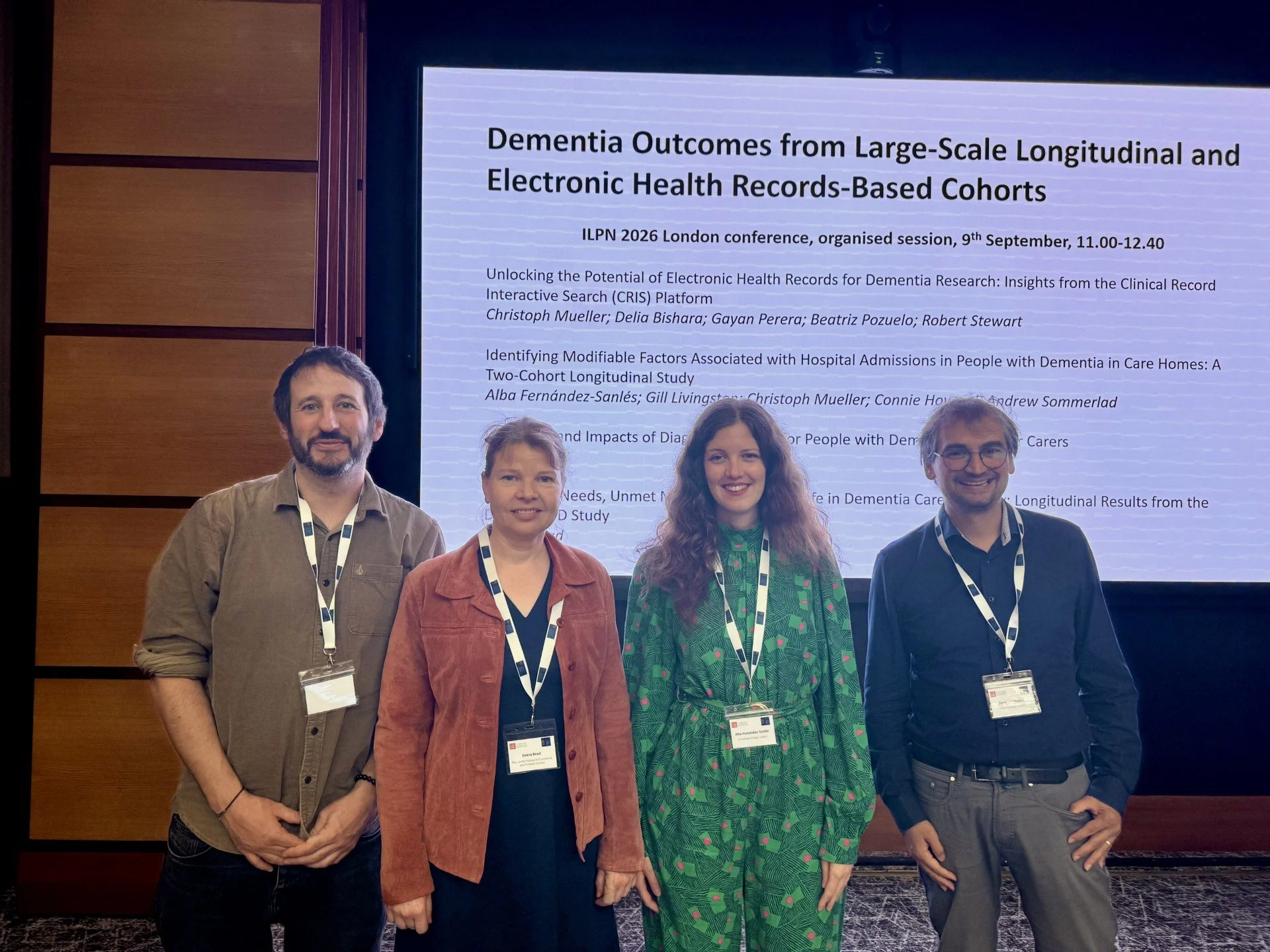 Four white researchers (two male, two female) standing in front of their presentation screen, smiling at the camera. They are all wearing white conference lanyards,.