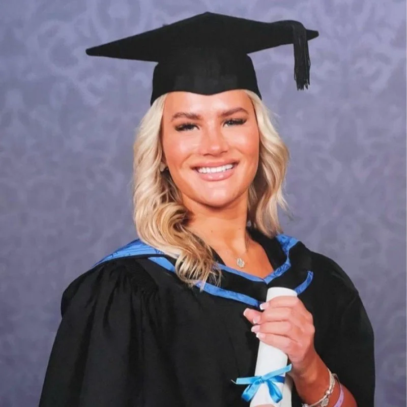 Woman in graduation gown and cap, holding diploma, smiling in front of a gray backdrop.