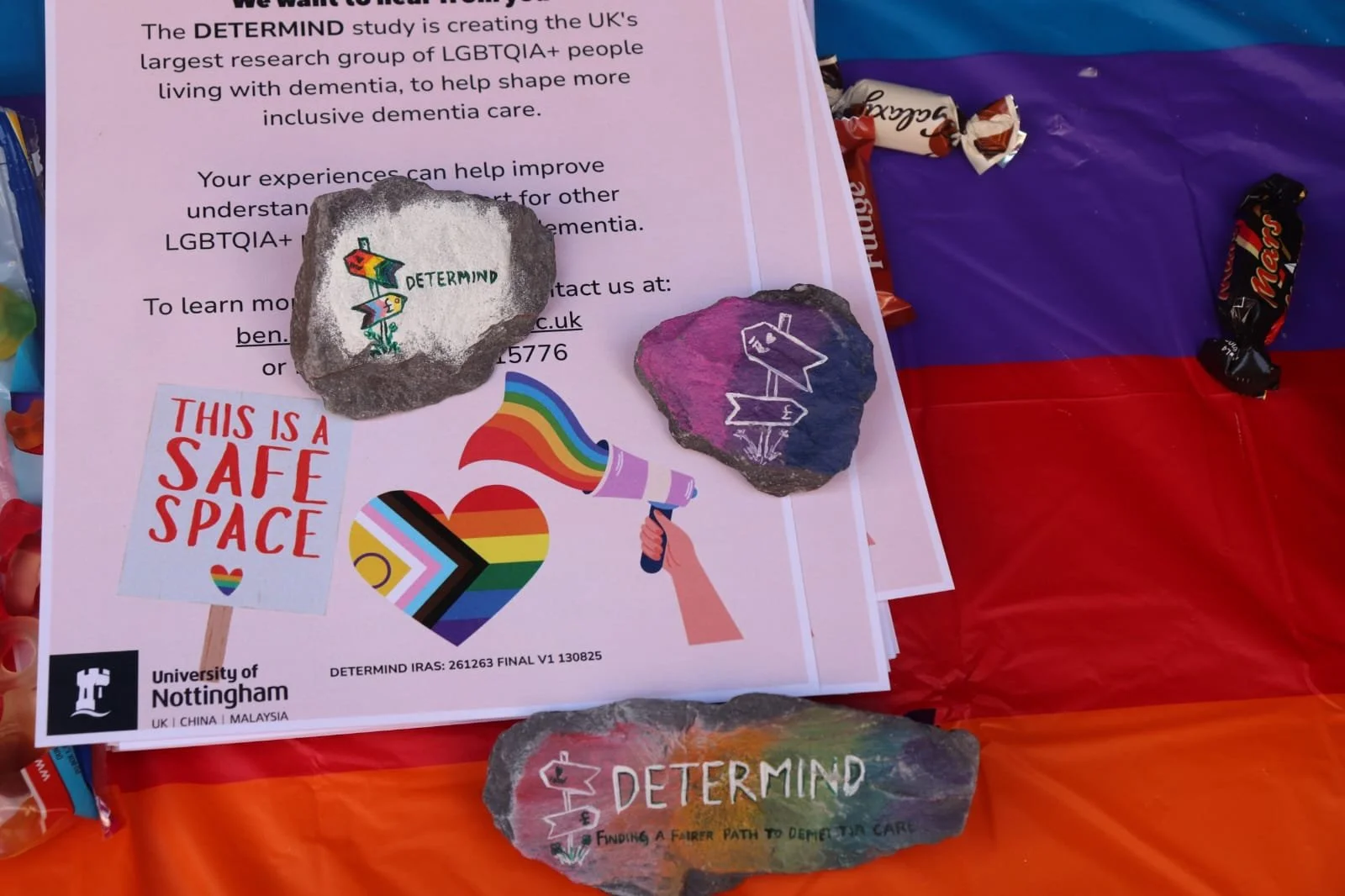 A close up of the DETERMIND leaflet, sitting on the pride stall table. It is surrounded by sweets and had two painted rocks on top of it. Some of the study team have painted the DETERMIND study logo against pride flag colours on the rocks.