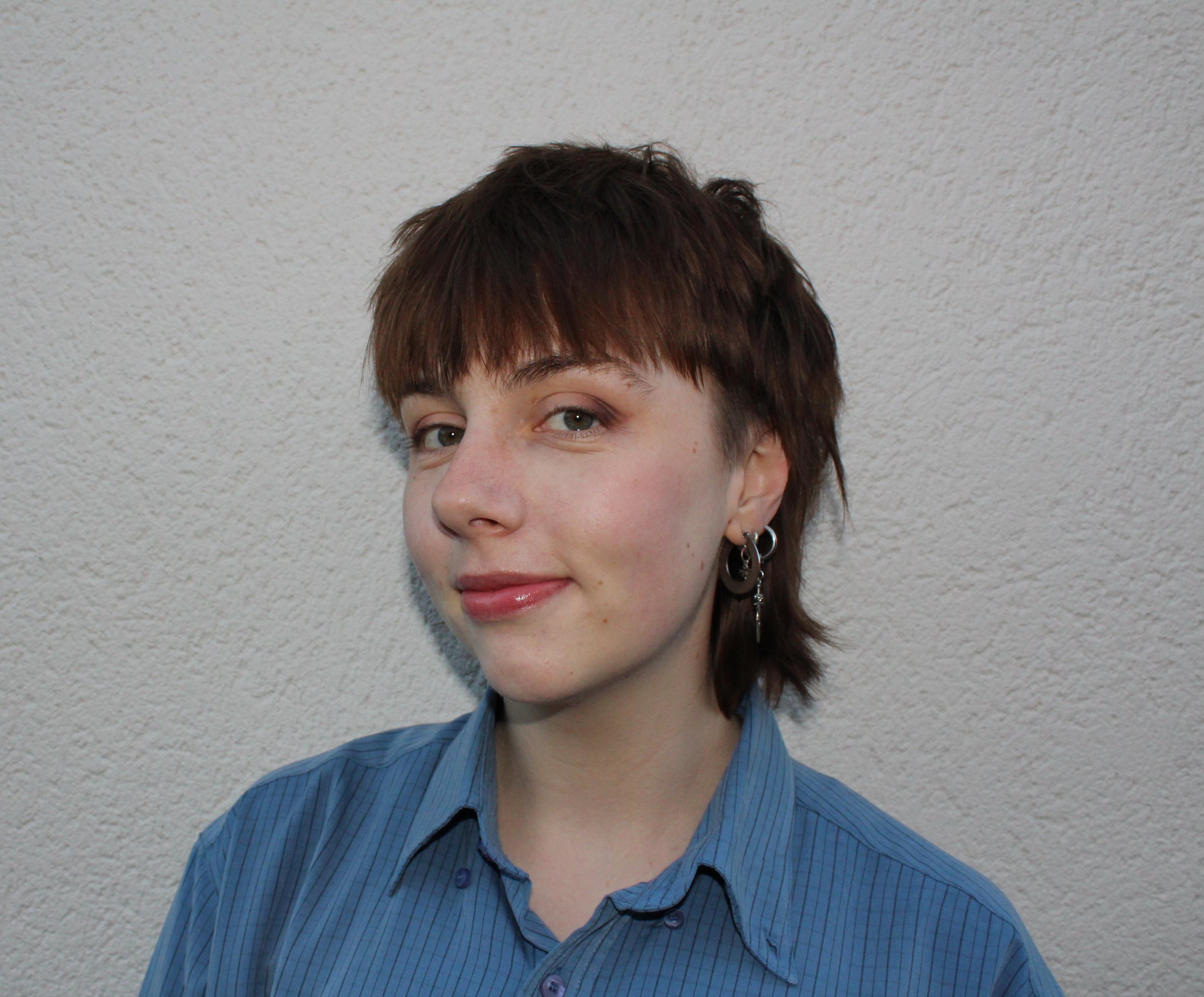 A person with brown hair standing in front of a plain background smiling. They are wearing a blue button up shirt