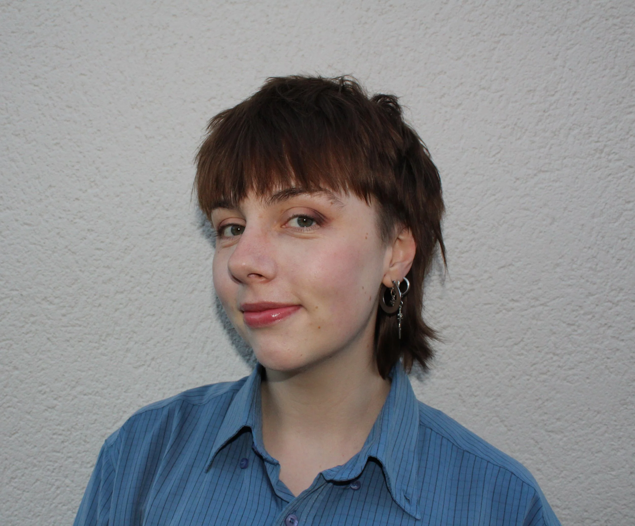 A person with brown hair standing in front of a plain background smiling. They are wearing a blue button up shirt