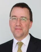 Portrait of a man with short dark hair, wearing glasses, a dark suit, white shirt, and a light-colored tie, against a light blue background.