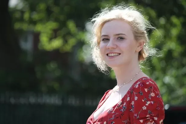 Woman with curly blonde hair wearing a red floral dress outdoors, smiling at the camera.