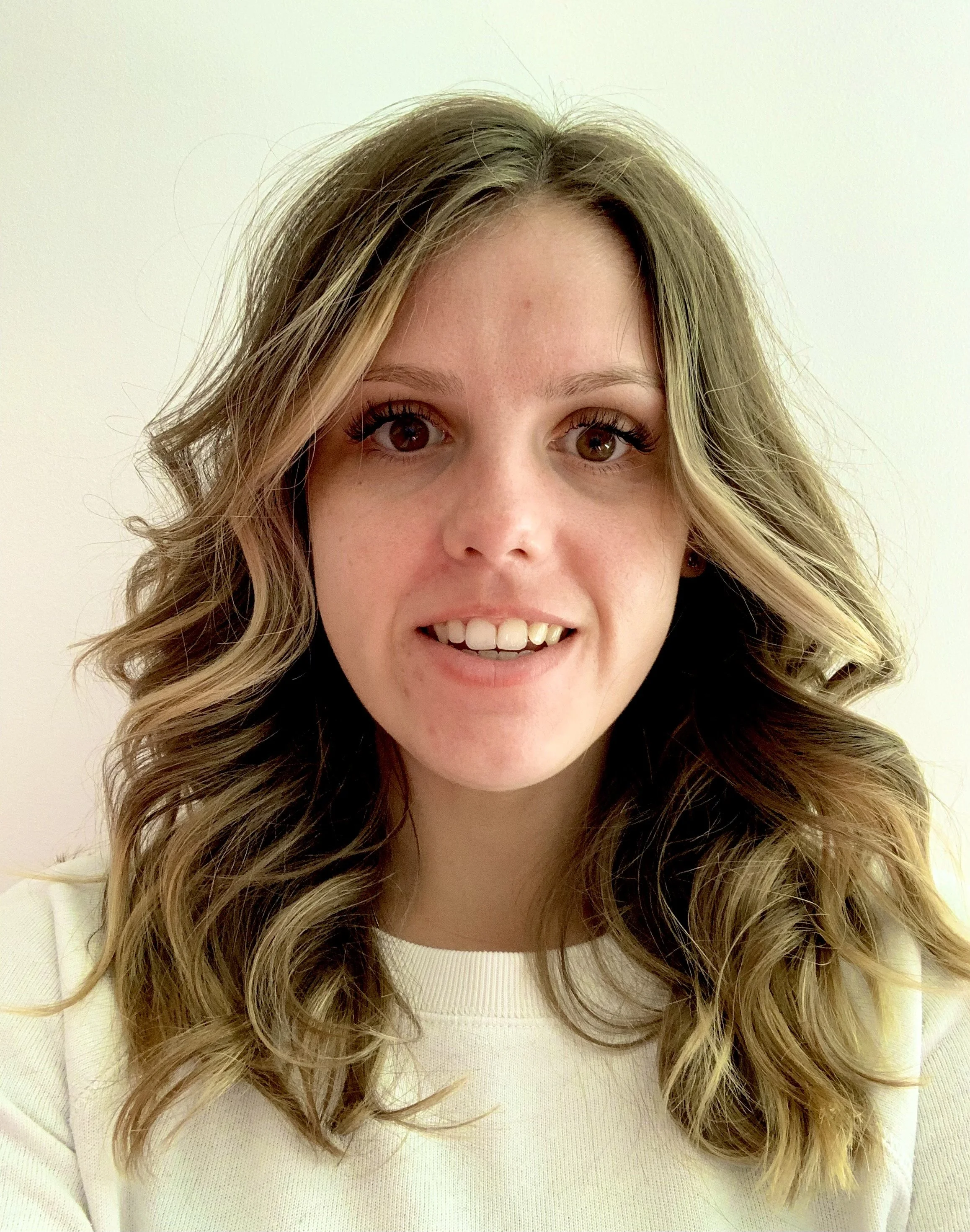 A woman with long brown hair wearing a white jumper. She is smiling at the camera.