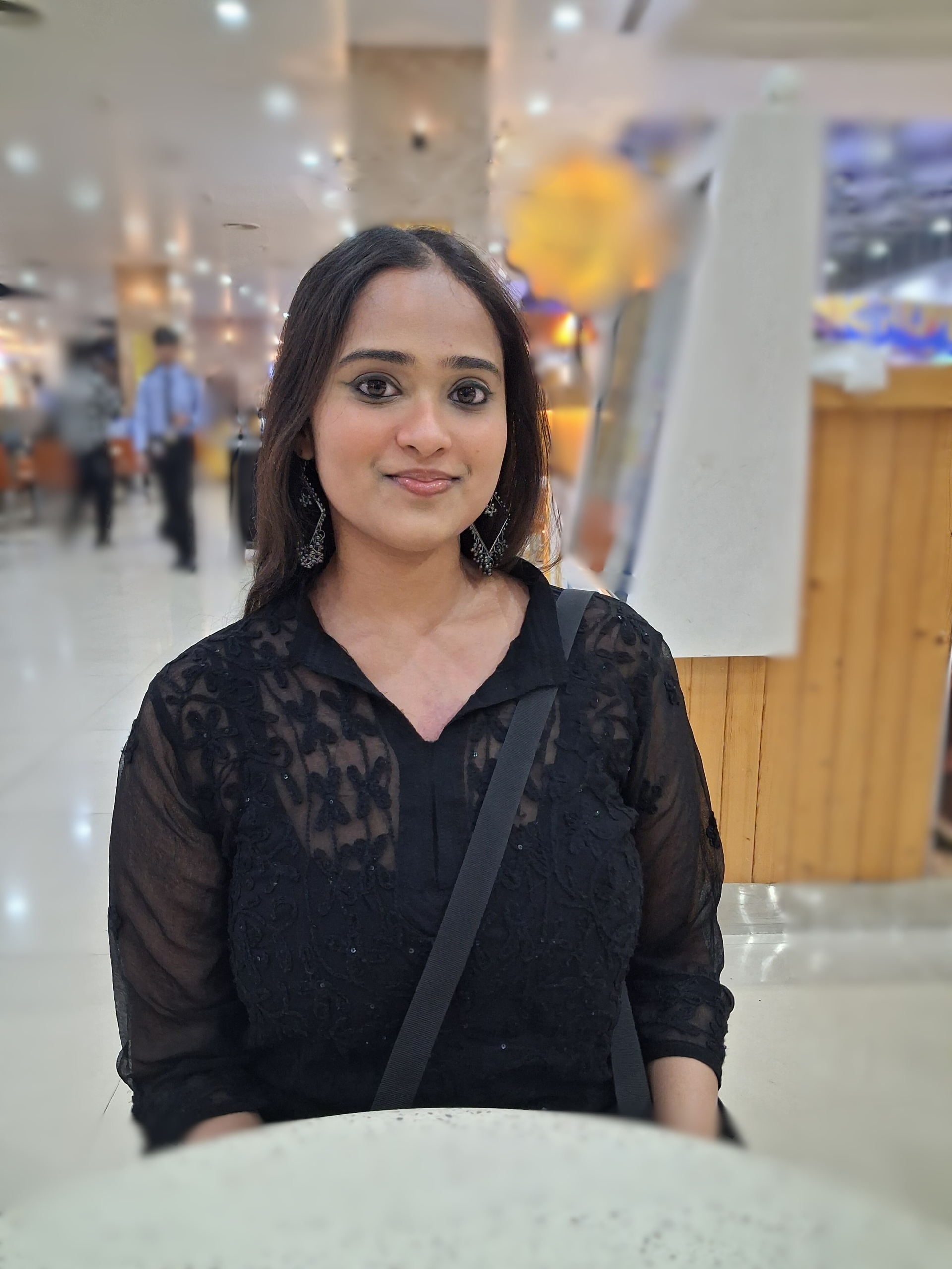 A person sitting at a table in a public place with the background blurred. She has long dark hair and is wearing a black button up shirt. She is smiling at the camera.